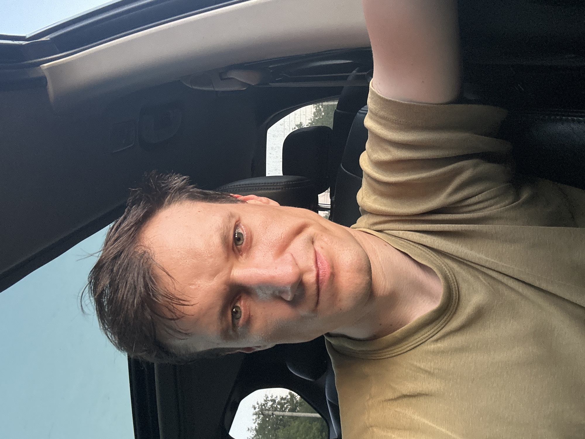 A man with short brown hair and green eyes is taking a selfie inside a car. He is wearing a light brown t-shirt. The car has a sunroof open, revealing a bright blue sky. Through the rear window, trees and foliage are visible, suggesting an outdoor location. The man is looking directly at the camera with a slight smile. The interior of the car appears to be dark, with black leather seats visible. The lighting suggests it might be late afternoon or early evening. The scene is consistent with a person taking a selfie while driving or stopped in their vehicle.