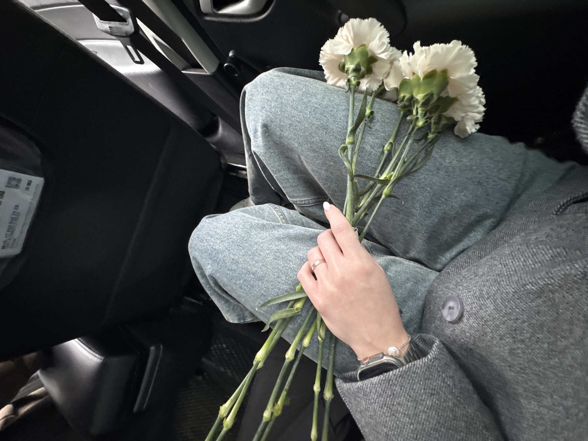 A person wearing light-wash denim jeans and a gray herringbone coat with a single button is holding a bouquet of white carnations. The person has a ring on their finger and a smartwatch with a pearl bracelet. The background shows the interior of a car with black seats and dashboard elements. The location is not discernible from the visual data, so it is impossible to determine if it is in Chaikovsky, Russia, or any other specific location.