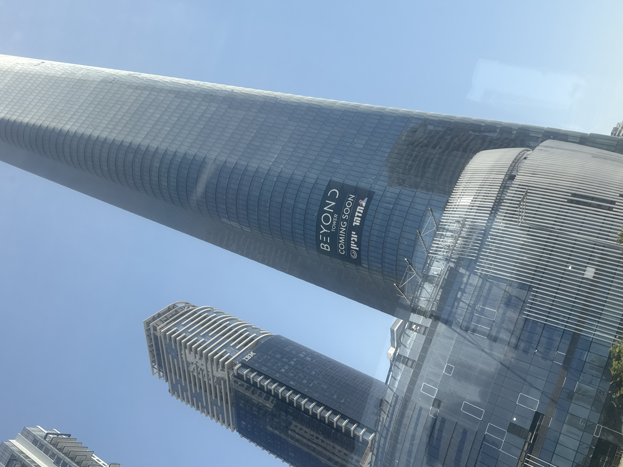 The image depicts several modern skyscrapers in Tel Aviv, Israel, against a clear, blue sky. One prominent, curved skyscraper is labeled "BEYOND TOWER COMING SOON". Another high-rise bears an "IBM" logo. All the structures are predominantly clad in reflective glass. The buildings are angled, as though the picture was taken from a moving vehicle.