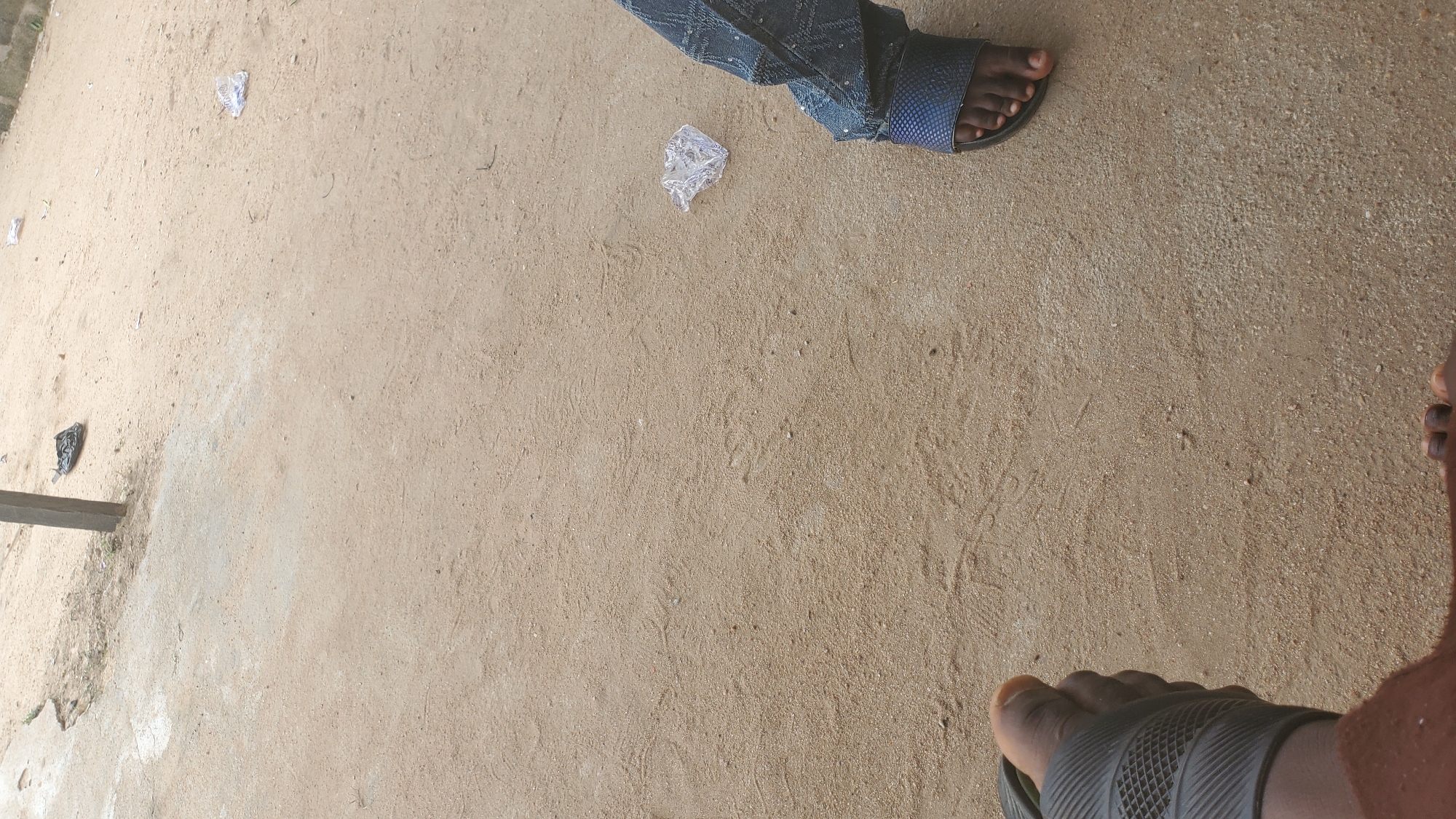 The image shows two individuals' feet on a sandy ground, likely in an outdoor urban setting. One person is wearing black sandals and has their right foot visible, with the toes pointing upwards. The other person is wearing blue patterned pants and dark blue sandals, with their right foot and leg clearly in view. The ground is composed of light brown sand with scattered debris, including a crumpled piece of white plastic. In the background, a vertical pole is visible, with a black plastic bag near its base. The scene suggests a casual outdoor gathering or resting spot, possibly in Jalingo, Nigeria, given the context.