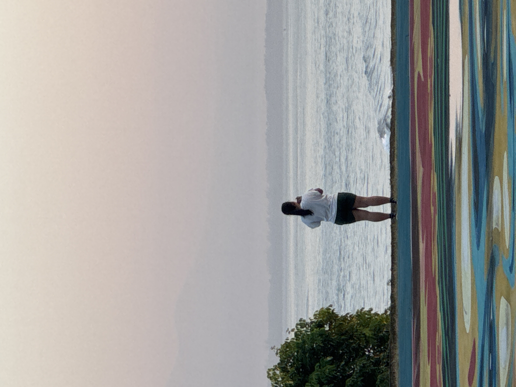 The image depicts a woman standing near the coastline in Kuta Selatan, Indonesia. She is wearing a white shirt and dark shorts and appears to be looking out at the water. The environment includes the ocean, a hazy sky, and a section of a colorful, vertical-striped wall. Some green vegetation is present at the bottom of the frame.