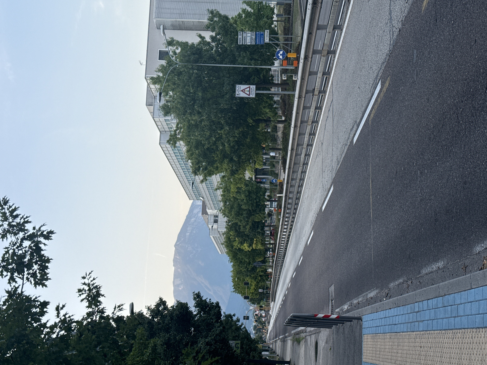 The image captures a street scene in Trento, Italy, with a mountain range visible in the background under a pale blue sky. The foreground is dominated by an asphalt road with white lane markings. To the left of the road is a pedestrian walkway with textured yellow paving and blue tiled sections. A metal guardrail separates the road from a sidewalk or green area on the right. Several trees with lush green foliage line the street, and a modern, multi-story building with glass windows is situated behind the trees. Traffic signals and road signs are present, indicating directions to various locations, including Trento, Magnete, Gardolo, and Canova. A few cars are parked or driving on the road in the distance. The overall impression is that of a well-developed urban area adjacent to a natural landscape.