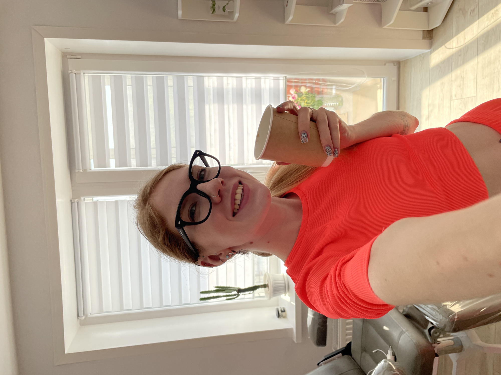 A woman wearing glasses and a bright orange cropped shirt is holding a brown paper cup in an indoor environment. She is smiling, and the cup is held near her face. Behind her, a window with closed white blinds is visible, along with some decorative items on the windowsill. The light suggests it may be daytime. A portion of an office chair is visible at the bottom of the image. There is nothing to indicate the location of Izhevsk, Russia.