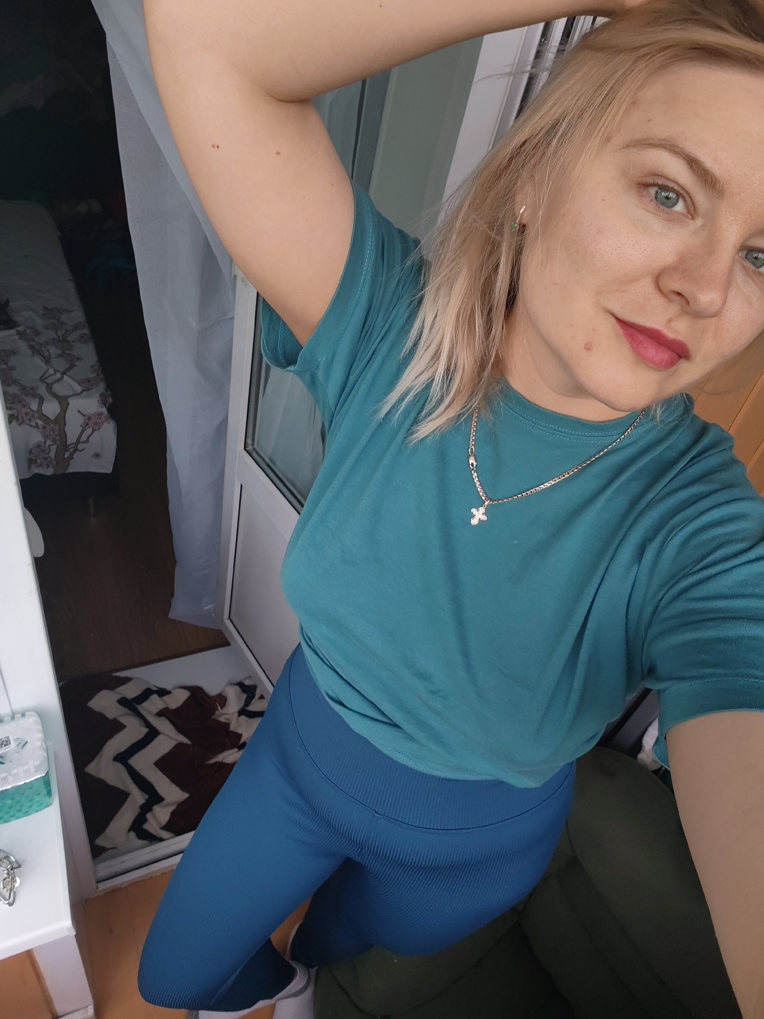 This is a close-up, high-angle selfie of a young woman with blonde hair and blue eyes, taken indoors. She is wearing a teal t-shirt and blue athletic leggings. A silver necklace with a small cross pendant is visible around her neck. Her left arm is raised behind her head, and she is looking at the camera with a slight smile.
The background shows a doorway leading to a room with a bed that has a floral-patterned duvet. A patterned rug is on the floor near the doorway. To the left, a white shelf holds a small green and white box and some keys. The lighting suggests it is daytime. The woman's attire and the presence of a rug and bed suggest a home or possibly a dorm room setting. The overall impression is casual and relaxed.