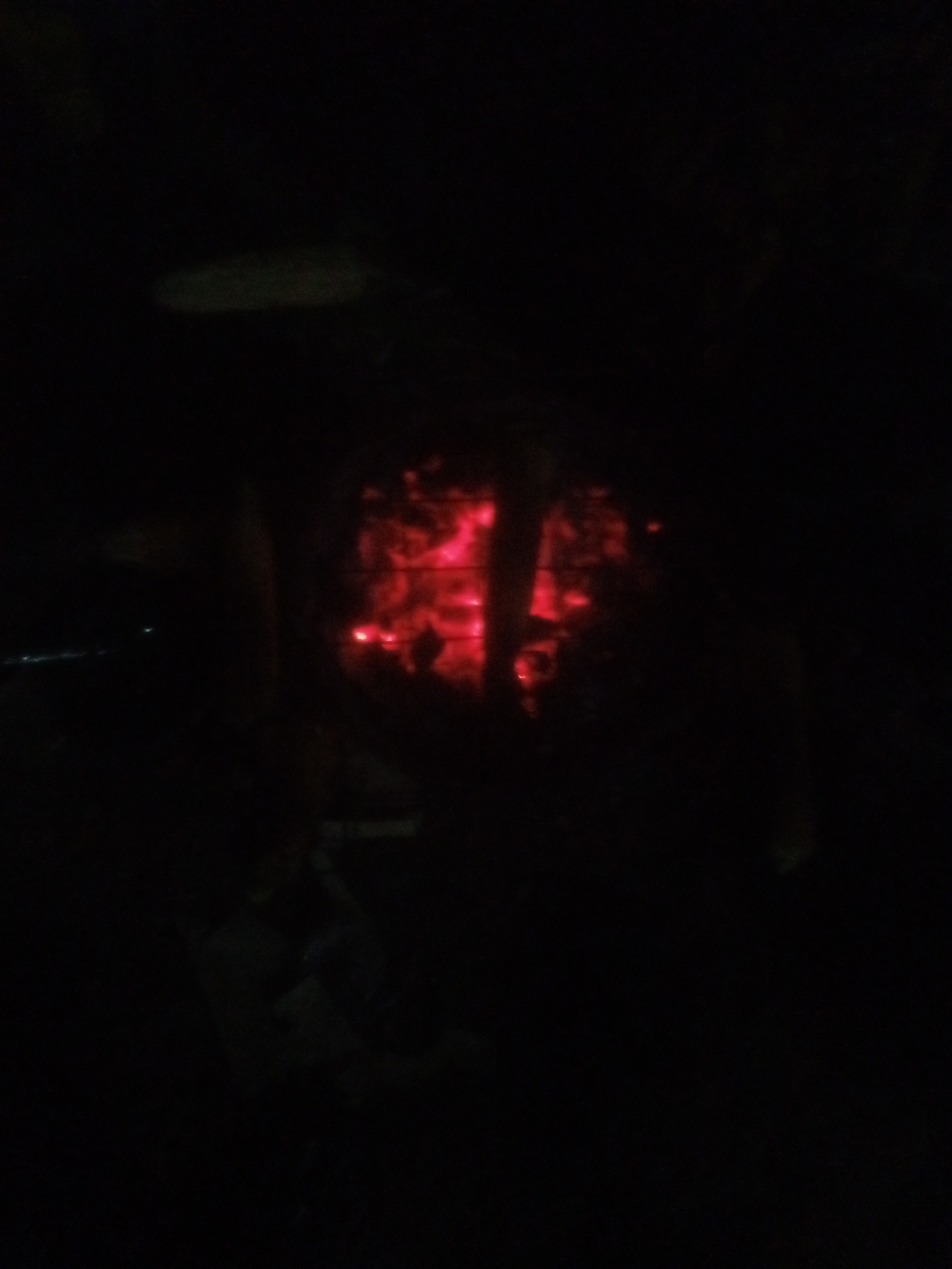 The image depicts a dimly lit scene, primarily composed of darkness. In the center, a glowing red area suggests the presence of fire or embers, possibly within a confined space. The red glow illuminates abstract shapes and textures, hinting at burning material. Surrounding this central focus is a very dark environment, with some faint, indistinct shapes visible in the background. There is a suggestion of an outdoor setting, possibly at night, with a faint horizontal line of lights in the far distance on the left, which could indicate a cityscape or road. The image quality is low, with significant noise and blur, making precise identification of objects and details difficult. The context of Tundun Wada West, Nigeria, suggests this could be a scene of a bonfire, cooking fire, or perhaps an incident involving fire within a residential or public area. The overall impression is one of a dark, ambient scene dominated by the intense red glow of heat and light.