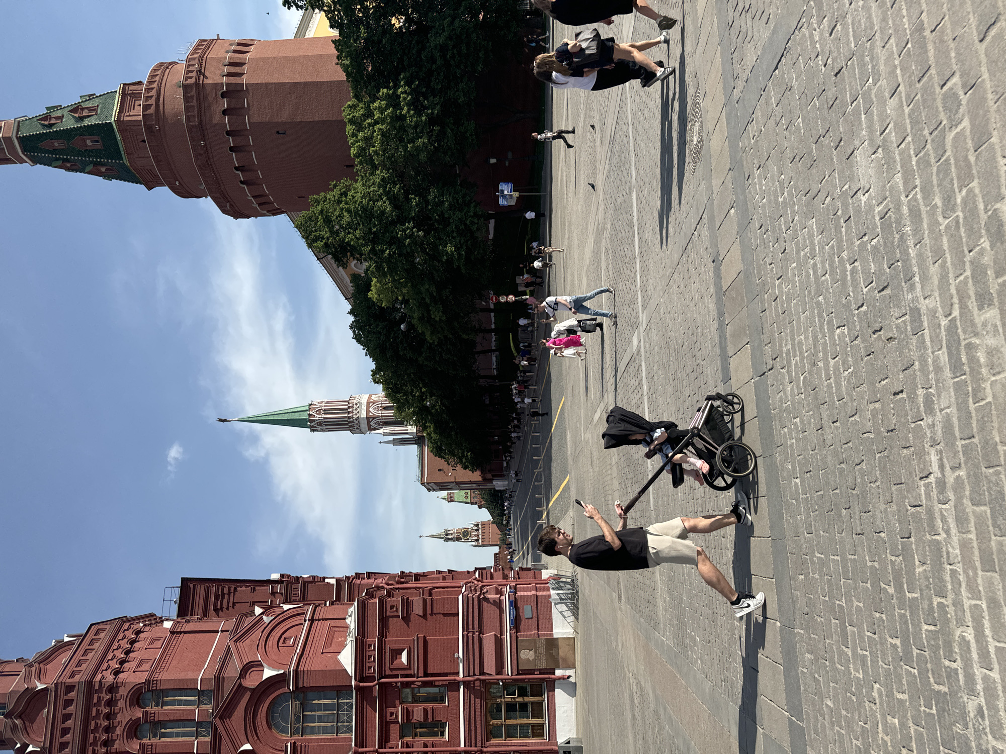 The image depicts a sunny day at Red Square in Moscow, Russia. A man in a black shirt and beige shorts is walking on the cobblestone pavement, pushing a baby stroller while looking at his phone. In the background are several iconic buildings: the State Historical Museum on the lower left, a series of Kremlin towers including the Spasskaya Tower, and trees lining the street. Other people are also seen walking in the distance.