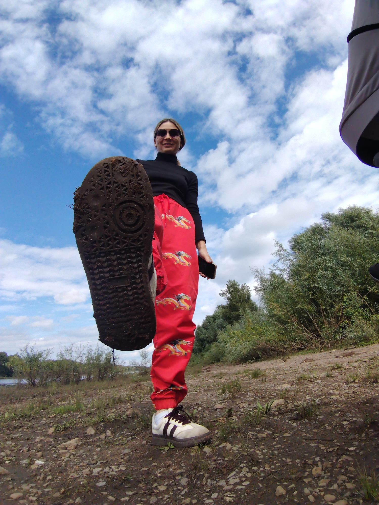 A low-angle, full-body shot captures a person standing outdoors, with their right foot raised and shoe sole facing the camera. The person is wearing a black turtleneck, red pants with a yellow dragon print, and white and black sneakers. They are also wearing sunglasses and holding a smartphone in their left hand. The background consists of a partially cloudy blue sky, lush green trees, and a body of water, suggesting a natural or semi-natural environment. The ground is covered with small rocks and sparse vegetation. The overall scene is a casual outdoor setting, possibly a park or waterfront area, with no immediate indication of the city of Chaikovsky, Russia.