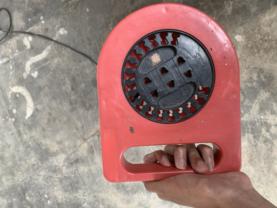 A person's hand holds a red plastic cable reel, commonly used as an extension cord holder, against a concrete background. The reel has a circular black panel at its center, featuring three horizontally aligned electrical sockets, likely Type C or F (two-pin), with a visible rating of "2.5A - 250V~". Below these sockets, the model number "EX-7013 P" is printed. To the right of the sockets, the "SNI" (Standard Nasional Indonesia) mark is clearly visible, confirming its compliance with Indonesian standards and indicating the location context of Metro, Indonesia. Above the main sockets, there's a small, recessed square-shaped element with internal markings that are difficult to discern but appear to be an indicator or symbol. The black central panel is surrounded by decorative, stylized patterns in red, integrated into the reel's design. The person's hand firmly grips a rectangular handle molded into the bottom of the red reel. The background is a grey concrete floor with scattered white paint splatters and a black electrical cable trailing from the left side of the frame, suggesting a utility or workshop setting. The image is lit by natural light, indicating it is daytime.