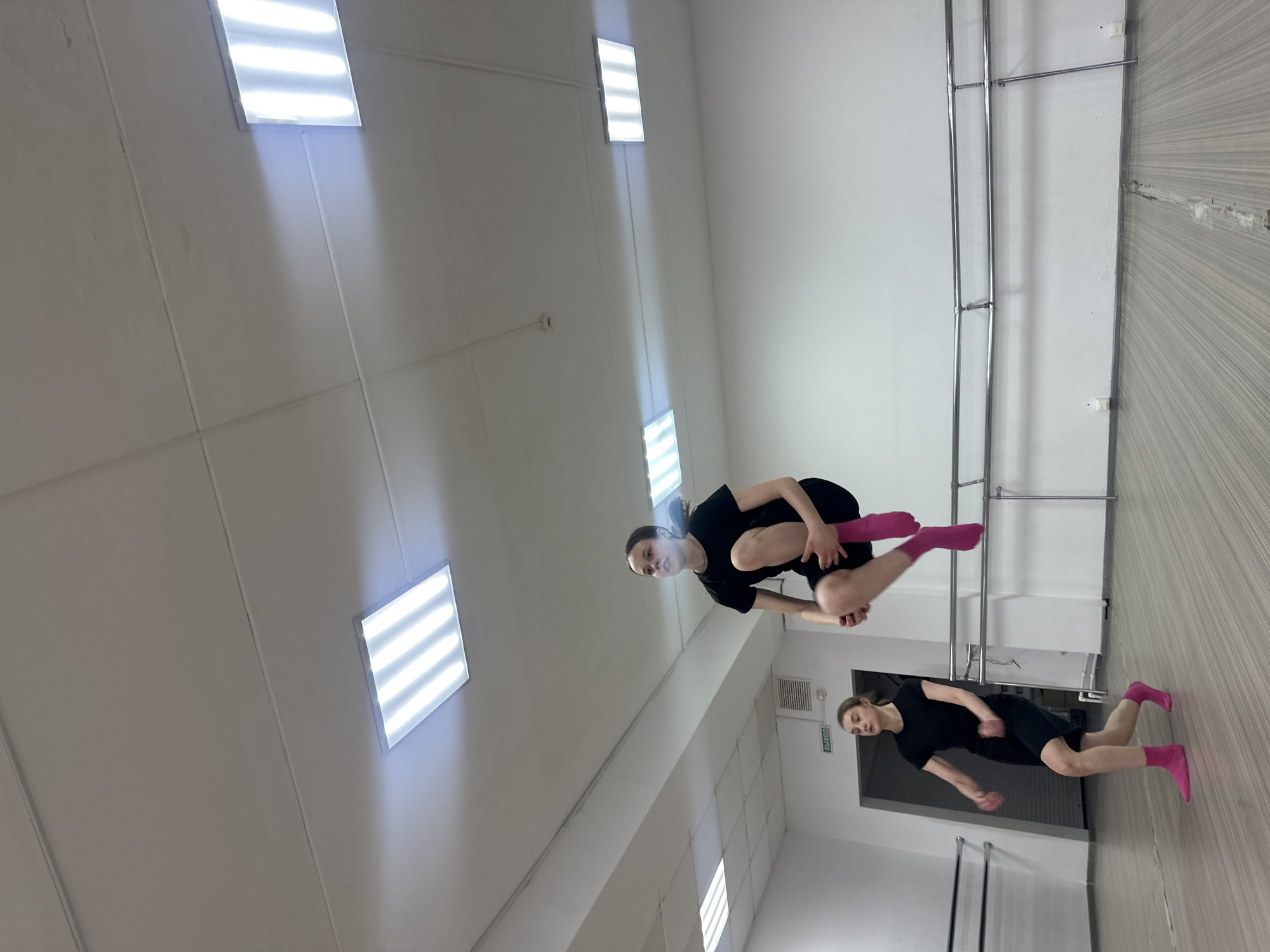 The image captures two young women engaged in what appears to be a dance or stretching session within an indoor studio located in Votkinsk, Russia.
The woman in the foreground is depicted in a dynamic pose, balancing on one leg (left) while her right leg is bent and lifted high, with the knee brought towards her chest. Her arms are bent, held close to her body, and she looks directly forward with a focused expression. The second woman is positioned further back and to the right, performing a lower, more grounded movement or stretch, with her arms slightly extended. Both women are wearing black short-sleeved tops, black shorts or skirts, and notable bright pink socks that cover their ankles and lower shins.
The setting is a well-lit studio. The ceiling, which appears to be composed of large white panels, features several rectangular fluorescent light fixtures emitting bright white light. These fixtures cast visible beams of light onto the adjacent surfaces. The walls are mostly white, contrasting with a section on the right side that is covered in light-colored, vertically planked wood. Along the white wall, parallel metal ballet barres are installed. The floor is made of light-toned wooden planks.
In the background, behind the second woman, a dark doorway is visible, above which a white sign with black Cyrillic text reads "ВЫХОД" (VYKHOD), meaning "EXIT". A ventilation grate is also present on the wall near the doorway. The overall impression is one of dedicated practice or rehearsal in a functional exercise space.