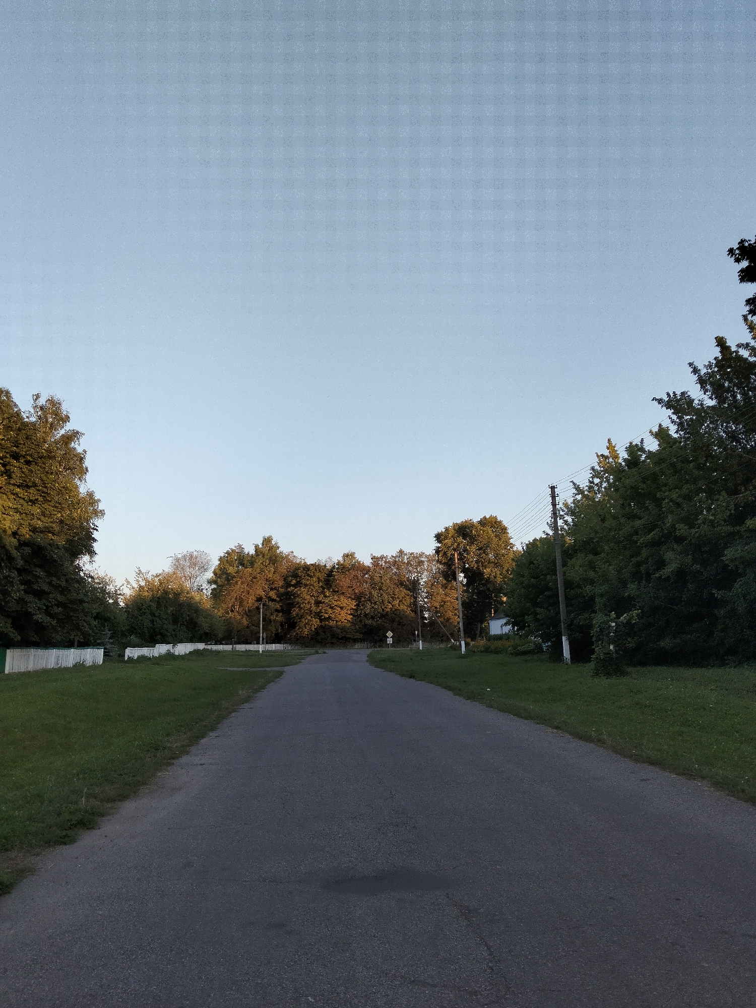A straight asphalt road stretches into the distance, flanked by green grass and trees on both sides. On the left, a white wooden fence runs along the edge of the grass. On the right, utility poles with power lines stand among the trees. The sky is clear and pale blue. The scene appears to be a rural road in Kliushnykivka, Ukraine, likely during the late afternoon or early evening, given the soft lighting.