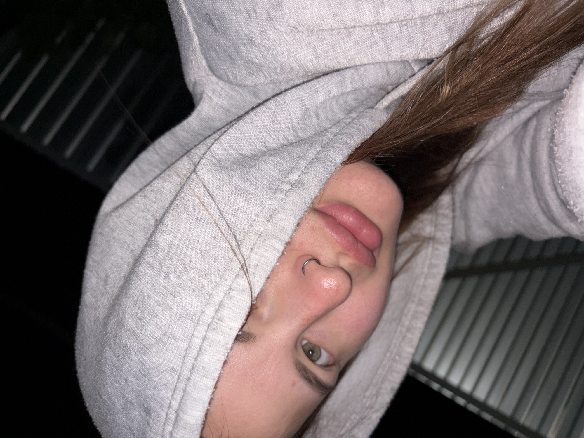 A close-up, slightly angled shot depicts a young woman wearing a light gray hooded sweatshirt. Part of her brown hair is visible beneath the hood. She has fair skin, a nose piercing, and green eyes. The background shows a dark area and a partially visible corrugated metal surface.
There is no specific information indicating this image was taken in Novopokrovskaya, Russia.
