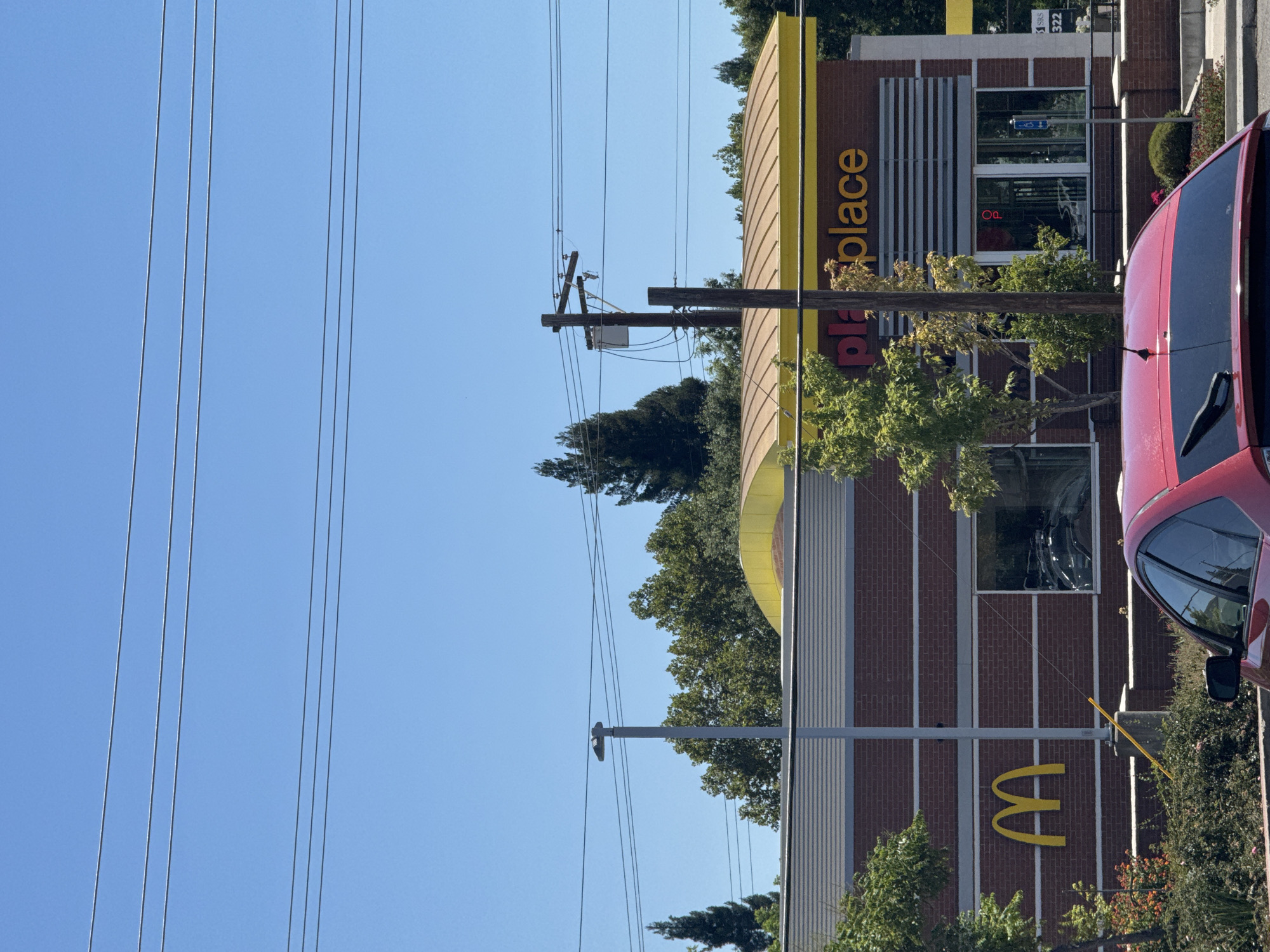 The image depicts a McDonald's restaurant in Citrus Heights, California. The building features a brick facade with a prominent yellow "M" logo on the left side. Above the entrance, the word "playplace" is visible in orange lettering. Power lines and utility poles are present in the foreground and background, with a clear blue sky overhead. A red car is parked in front of the establishment, partially obscuring the lower portion of the building. Trees and landscaping surround the building and parking area. The overall scene suggests a typical sunny day in a suburban commercial area.