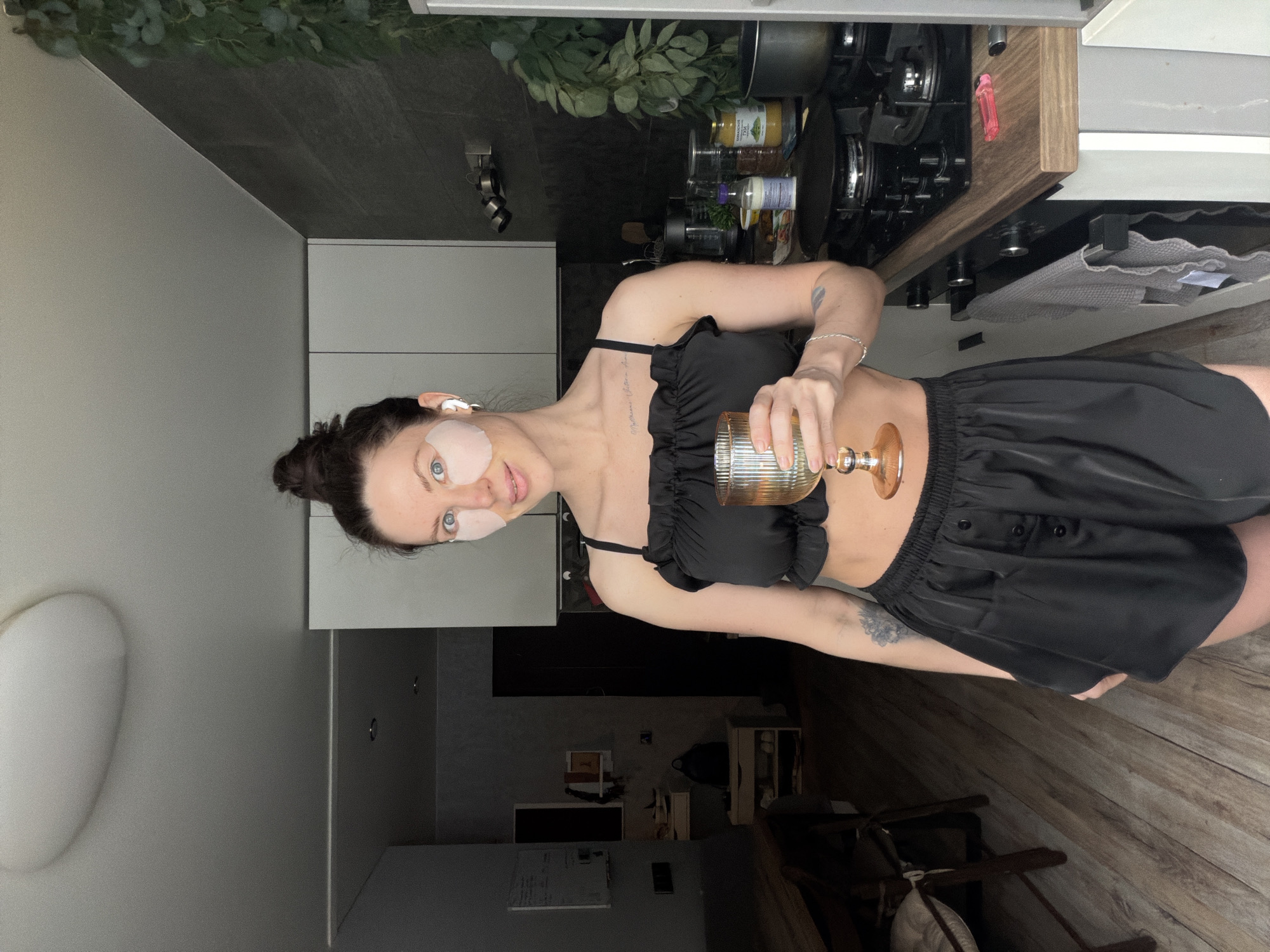 A woman is captured in an indoor setting, appearing to be in a modern kitchen and living area, located in Perm, Russia. She is wearing a black, ruffled crop top and black shorts. Her dark hair is styled in a bun, and she has light-colored under-eye patches applied to her face, suggesting a self-care or relaxed moment. She is holding a clear, ribbed glass with a golden stem and rim in her right hand, possibly containing a drink.
The woman looks directly at the camera with a neutral to slightly smiling expression. Visible tattoos include a design on her left forearm and text on her collarbone that reads "Memento vivere".
The background reveals a well-equipped kitchen to her right, featuring a stovetop, various cooking utensils, and modern cabinets. Above the stovetop, there are decorative green plants or foliage. Several jars and bottles are visible on the counter, with text labels such as "ГОРЧИЦА СЛАДКАЯ" (Sweet Mustard), "Майонез" (Mayonnaise), and "СОВУШКА" (Sovushka - Little Owl) on a clear bottle. To her left and further back, a living space is discernible with dark furniture, possibly a television or monitor, and chairs. The floor throughout the visible area is made of wooden laminate. The lighting suggests daytime indoors.