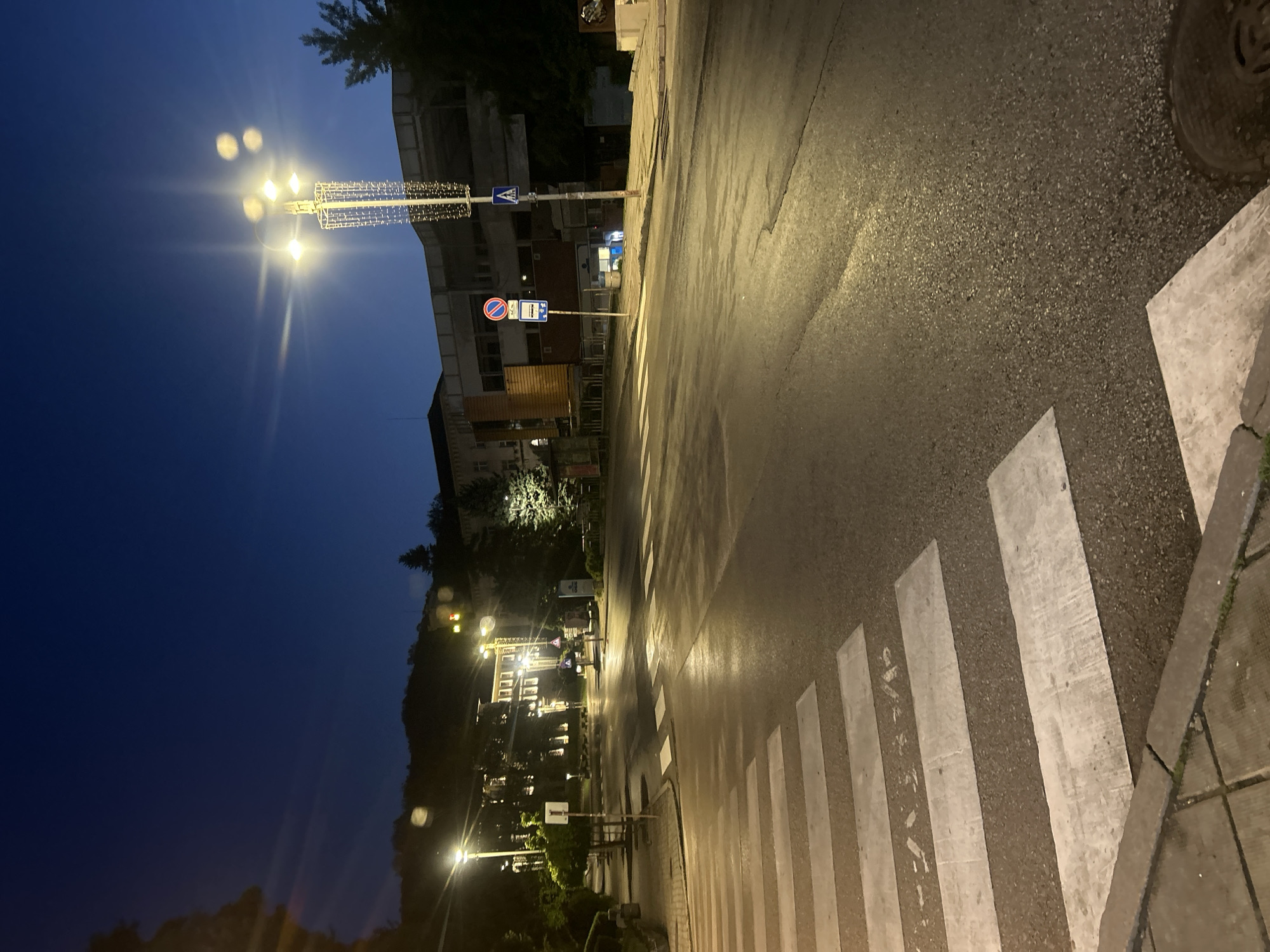 The image depicts a street scene at night in Veliko Tarnovo, Bulgaria. A pedestrian crosswalk is prominent in the foreground, illuminated by streetlights. The road surface appears wet, reflecting the ambient light. Buildings line the street, their architecture characteristic of the city. Street signs are visible, indicating local traffic regulations. Streetlights with decorative elements are present, adding to the ambiance. The sky is dark, suggesting the image was taken after sunset. No people are visible in the frame.