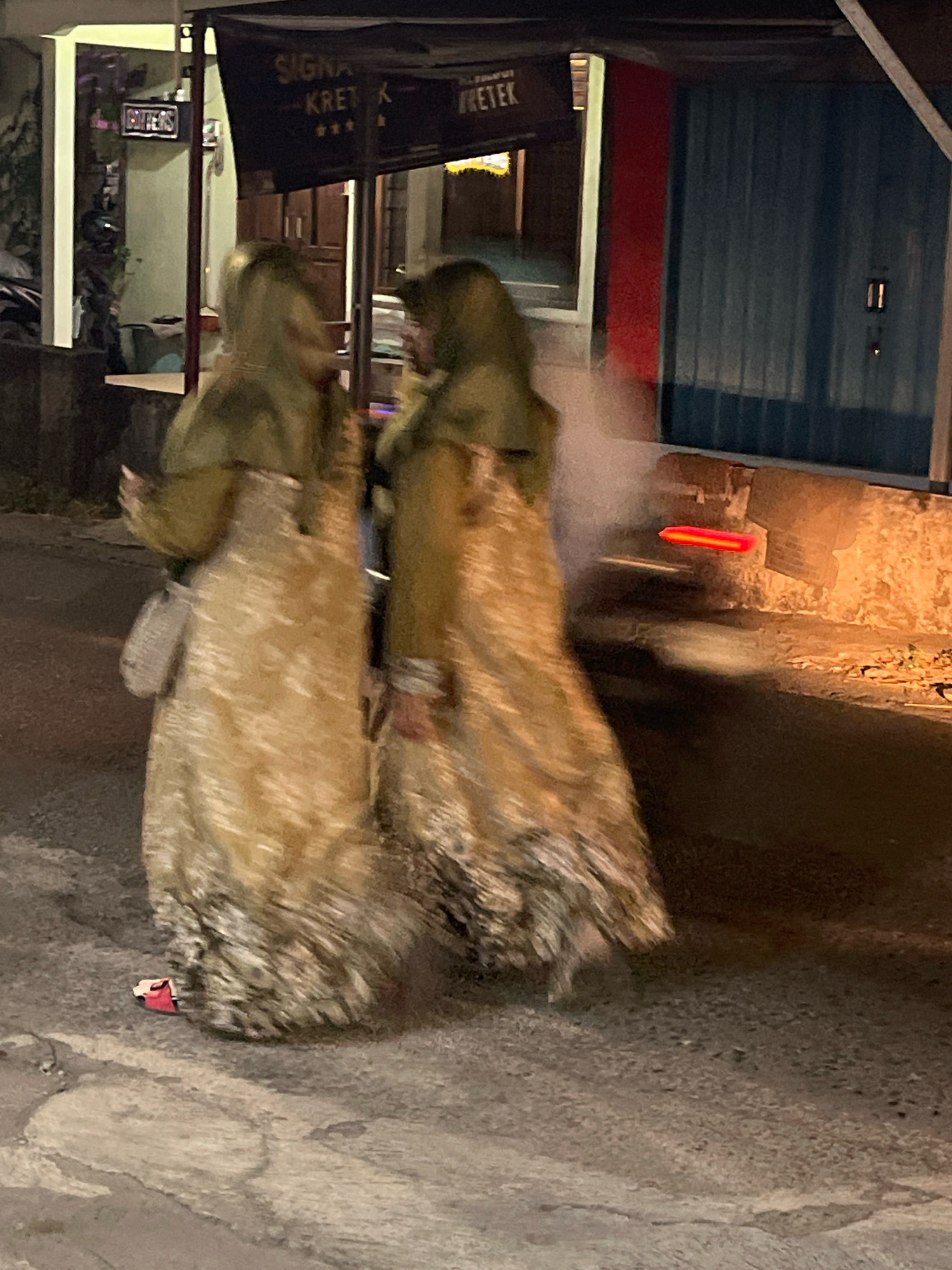 Two individuals, wearing hooded, floor-length garments with a patterned texture, are walking on a paved street at night. The person on the left carries a light-colored bag over their shoulder. A small, red object, possibly footwear, is visible on the ground near the feet of the person on the left. In the background, a building with a dark awning displays text that includes "SIGNATURE KRET K" and "RETEK". To the left of the awning, a sign with the word "GRITTERS" is illuminated. The environment appears to be an urban setting in Banguntapan, Indonesia, with a brick wall and a blue corrugated metal structure visible in the distance. There is a bright, horizontal red light source in the mid-ground, suggesting illumination from a vehicle or signage.
