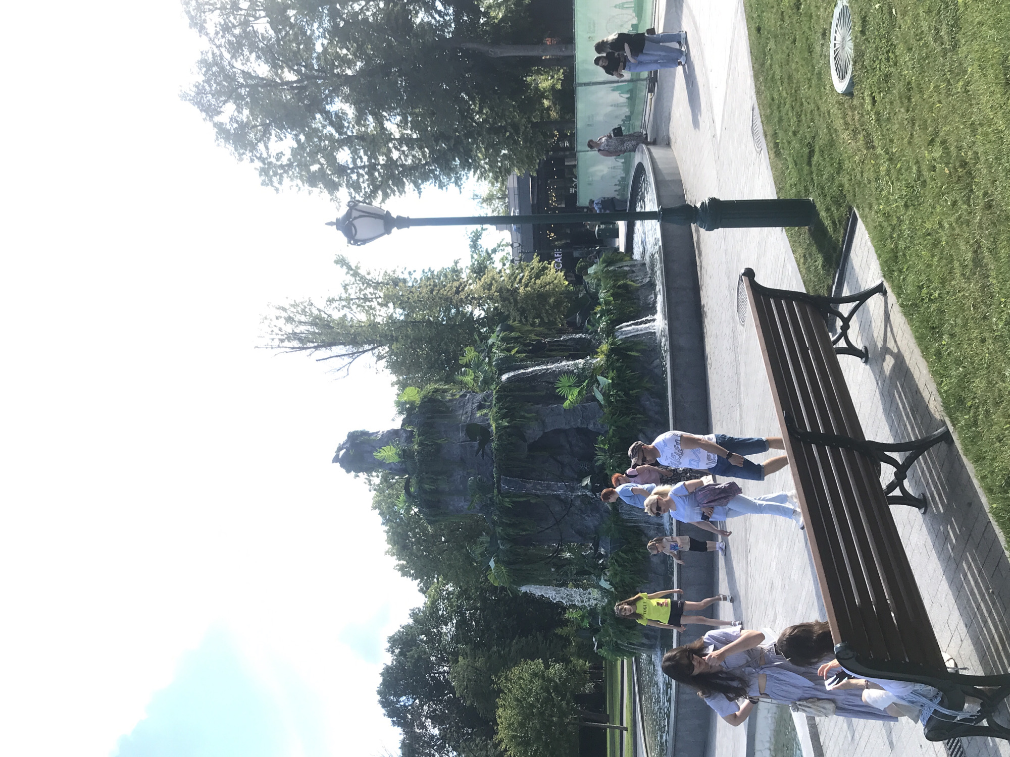 A group of people are walking around a multi-tiered fountain decorated with artificial rocks and lush greenery. The fountain is the central focus of the image, with water cascading down its sides. A park bench sits in the foreground, casting shadows on the grass. A lamppost stands to the right of the fountain, and trees form a backdrop. The sky is bright, suggesting a sunny day.