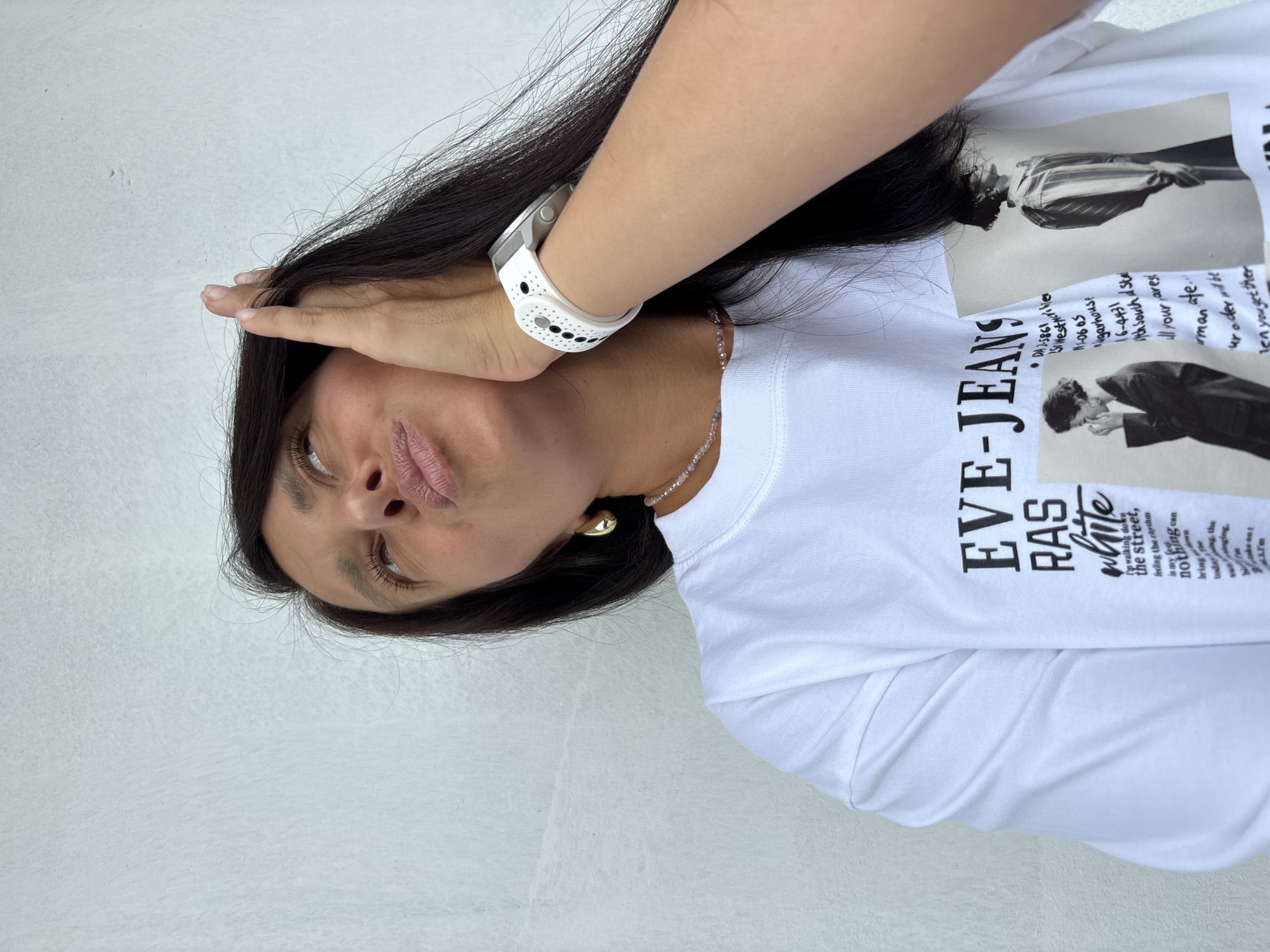 A woman with long, dark hair is captured in a close-up, angled shot against a white, textured wall. She is wearing a white t-shirt with a graphic print that includes text and two black and white images of people. Around her neck, she has a delicate beaded necklace, and a gold hoop earring is visible on her left ear. On her left wrist, she wears a white digital watch with a perforated strap. Her right hand is raised, with fingers gently touching her hair near her temple, and her head is tilted back slightly, with her gaze directed upwards. Her lips are pursed. The lighting suggests an outdoor setting, likely during the daytime. There is no indication of the location being in Chaikovsky, Russia.