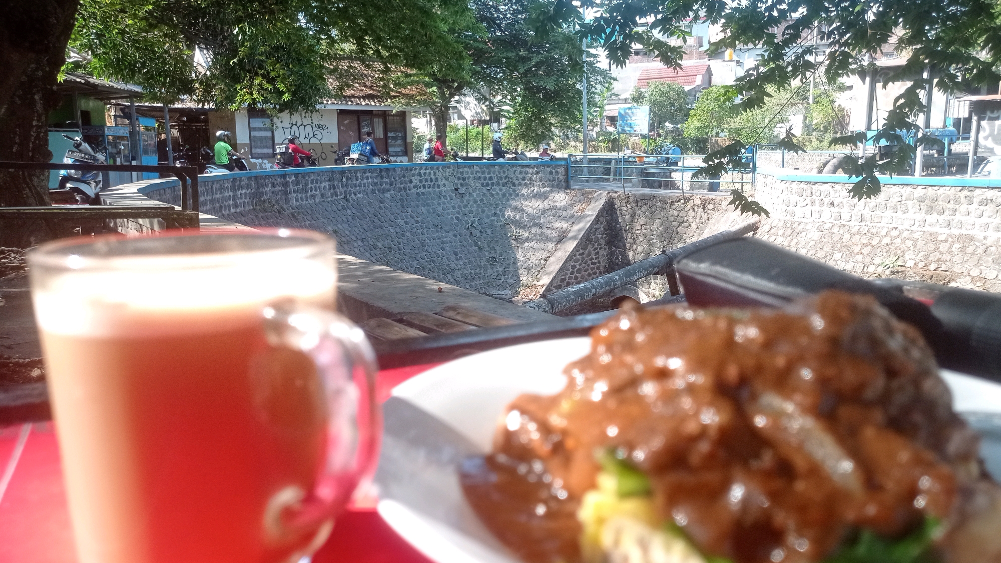 In Mulyoagung, Indonesia, a close-up shot focuses on a mug of beverage and a plate of food. The beverage, a light brown color, is in a clear mug with a handle, positioned on the left side of the frame. To the right, a plate of food is visible, featuring what appears to be meat in a rich, dark brown sauce. The background reveals an outdoor setting with a stone-lined canal or drainage ditch. Beyond the canal, several buildings and trees are visible under a bright, sunny sky. Motorcycles are parked and in use along a walkway bordering the canal, with some individuals on them. The overall scene suggests a casual outdoor dining or resting spot with a view of the local environment.