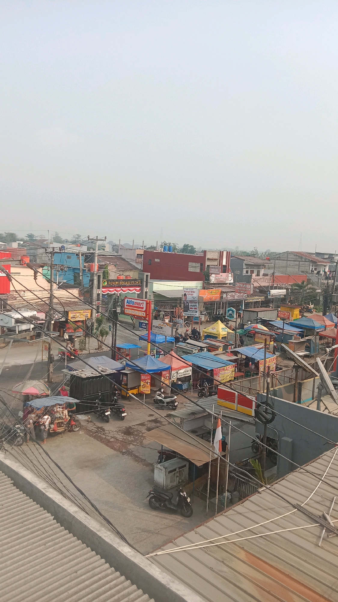 This is an eye-level, medium shot of a street scene in Jalenjaya, Indonesia, with an overcast sky. The foreground shows a textured, corrugated metal roof. The mid-ground is a busy street with numerous small food stalls set up under colorful awnings, some with people standing or sitting nearby. Various motorcycles are parked or in use on the street, which appears to be paved with asphalt and some dirt. Electrical wires are prominently strung across the scene, supported by utility poles. In the background, buildings of varying sizes and colors, including what appears to be a commercial building with an "Alfamart" sign, are visible. A few larger buildings with red roofs are also present. The overall impression is of a bustling, informal commercial area.