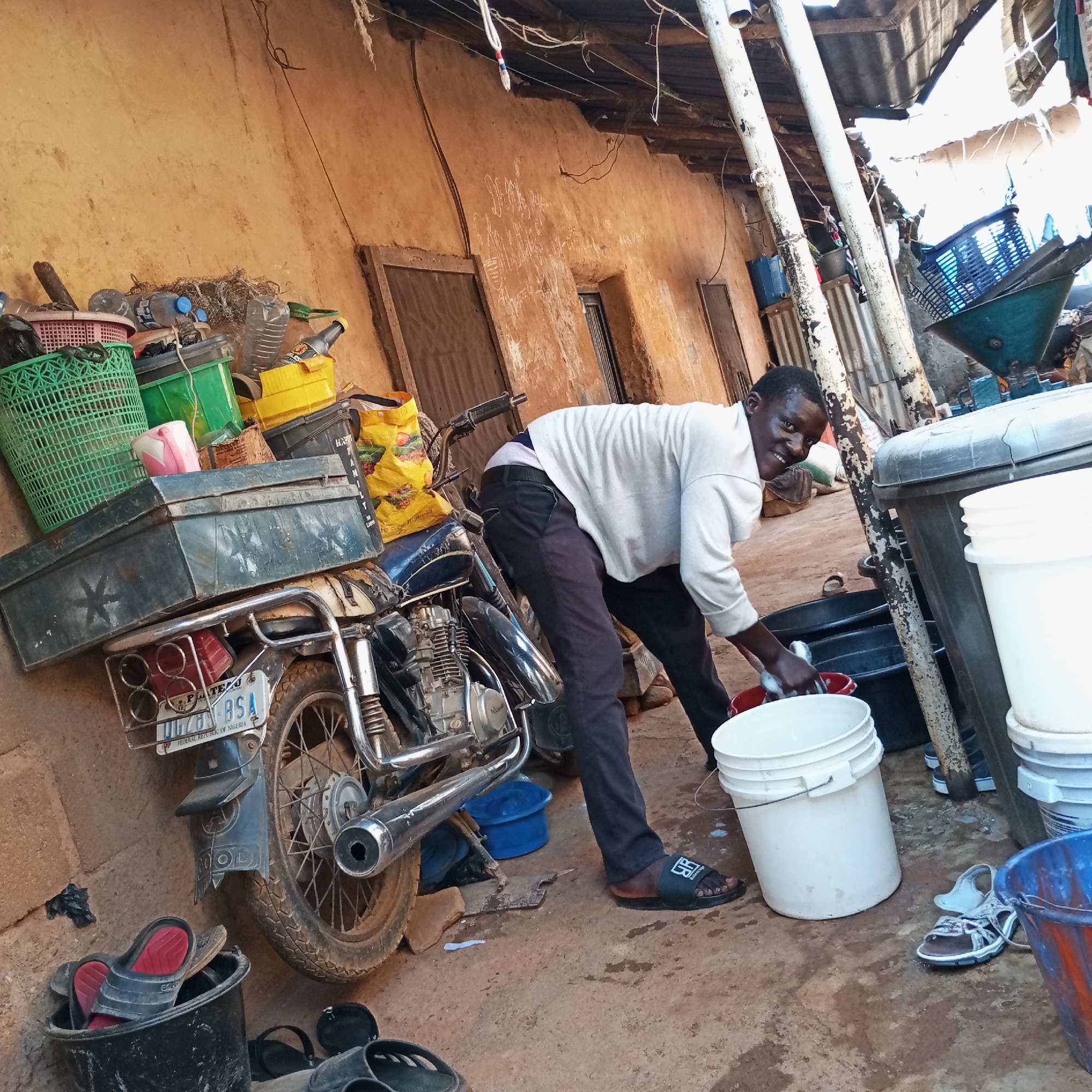A man is washing dishes outdoors in Jos, Nigeria. He is wearing a white long-sleeved shirt, dark pants, and black sandals. He is leaning over a collection of buckets and tubs, washing a white dish in a red tub. To his left, a motorcycle is parked against a mud wall, loaded with baskets and bags. The motorcycle license plate reads “Plateau” and "UG-785 BSA". The background shows a dilapidated building with a corrugated metal roof. The ground is dirt. Other buckets and shoes are scattered around the area.