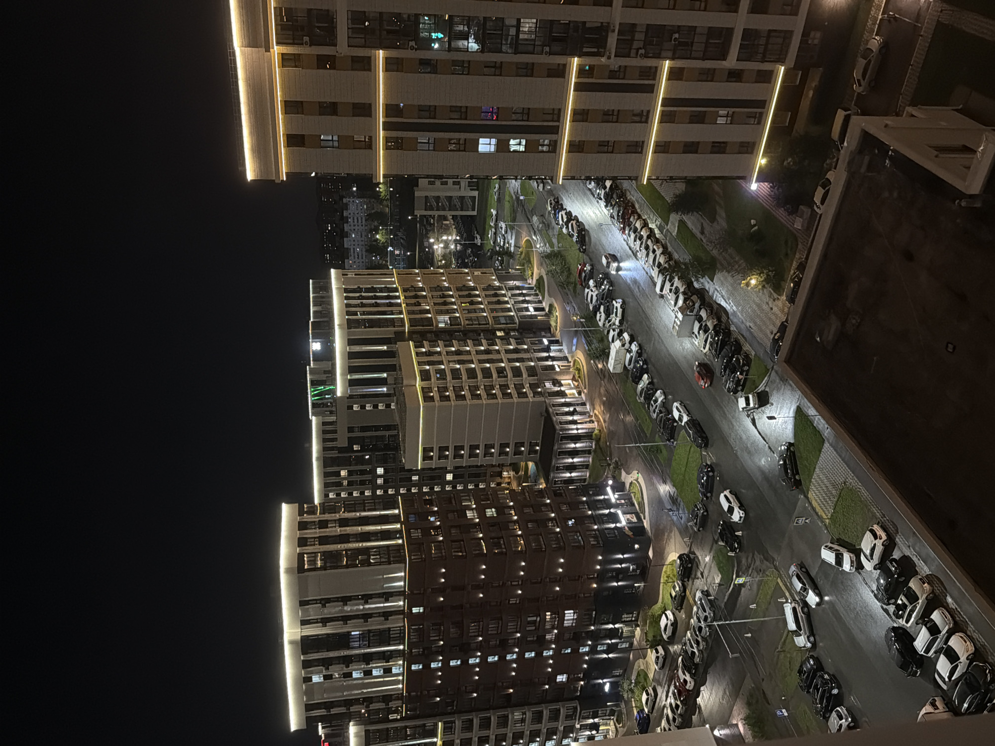 The image shows a night view of a modern residential complex in Barnaul, Russia. Several multi-story apartment buildings with illuminated facades and windows are visible. A street lined with parked cars runs through the complex, with streetlights casting pools of light on the wet asphalt. The overall impression is one of urban living at night, with the lights of the buildings and cars creating a vibrant, yet tranquil, atmosphere.