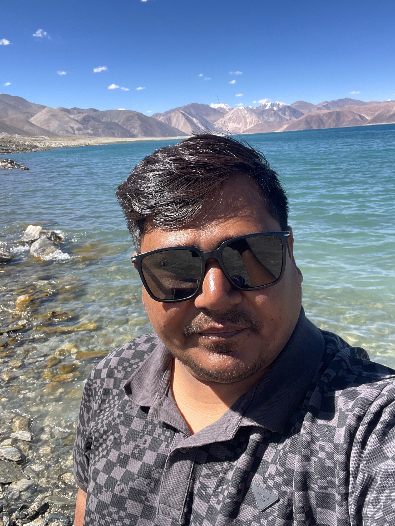 The image is a selfie taken by a man wearing sunglasses and a grey checkered polo shirt. He is standing by a body of water with rocky shores and mountains in the background. The water is a clear turquoise color, and the sky is blue with a few white clouds. The mountains are rugged and appear to be snow-capped in some areas. The scene suggests a natural, possibly remote, location. The man's attire and the sunglasses indicate a sunny day. The overall impression is one of a scenic travel photo.