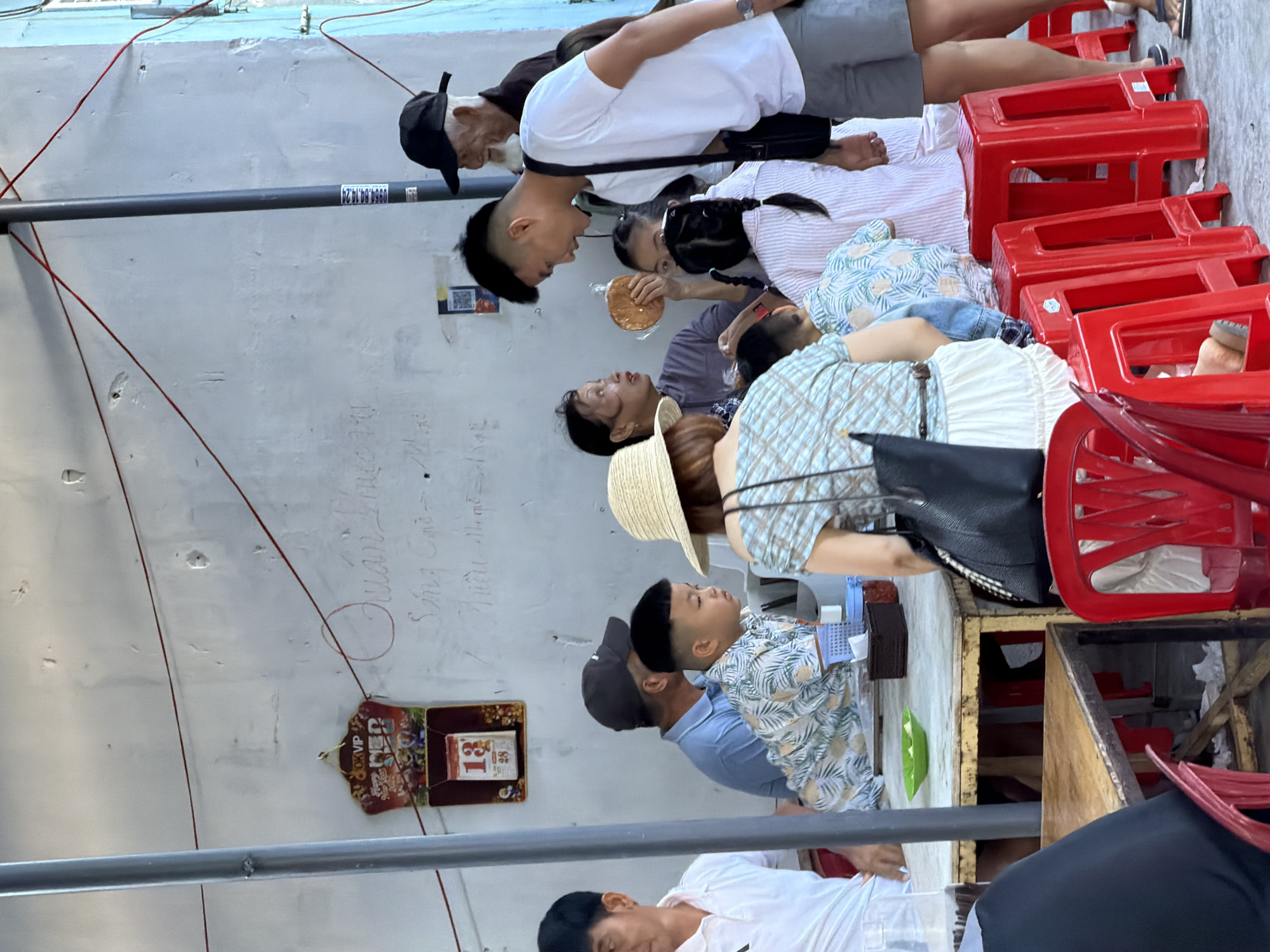 A group of people are gathered around tables outside. The scene appears to be a food stall or casual dining area in Bac Nha Trang Ward, Vietnam. In the foreground, a man in a white shirt and gray shorts is standing, facing away from the camera. To his left, a woman in a patterned shirt and white skirt is seated, also facing away. In the middle ground, several individuals are seated around a table. A young boy in a patterned shirt is looking to the right. An adult male wearing a blue shirt and a dark cap is behind him. A woman with a straw hat and a purple shirt is seated facing the boy. Another woman, holding a round, flat bread-like object, is speaking to a young girl with braided hair. An older man with a white beard and a black cap is standing behind the group, to the right. A calendar with the number 13 prominently displayed hangs on the wall behind the table on the left. The environment consists of a rough, white-painted wall, red plastic stools, and wooden tables. Red wires are strung across the wall.