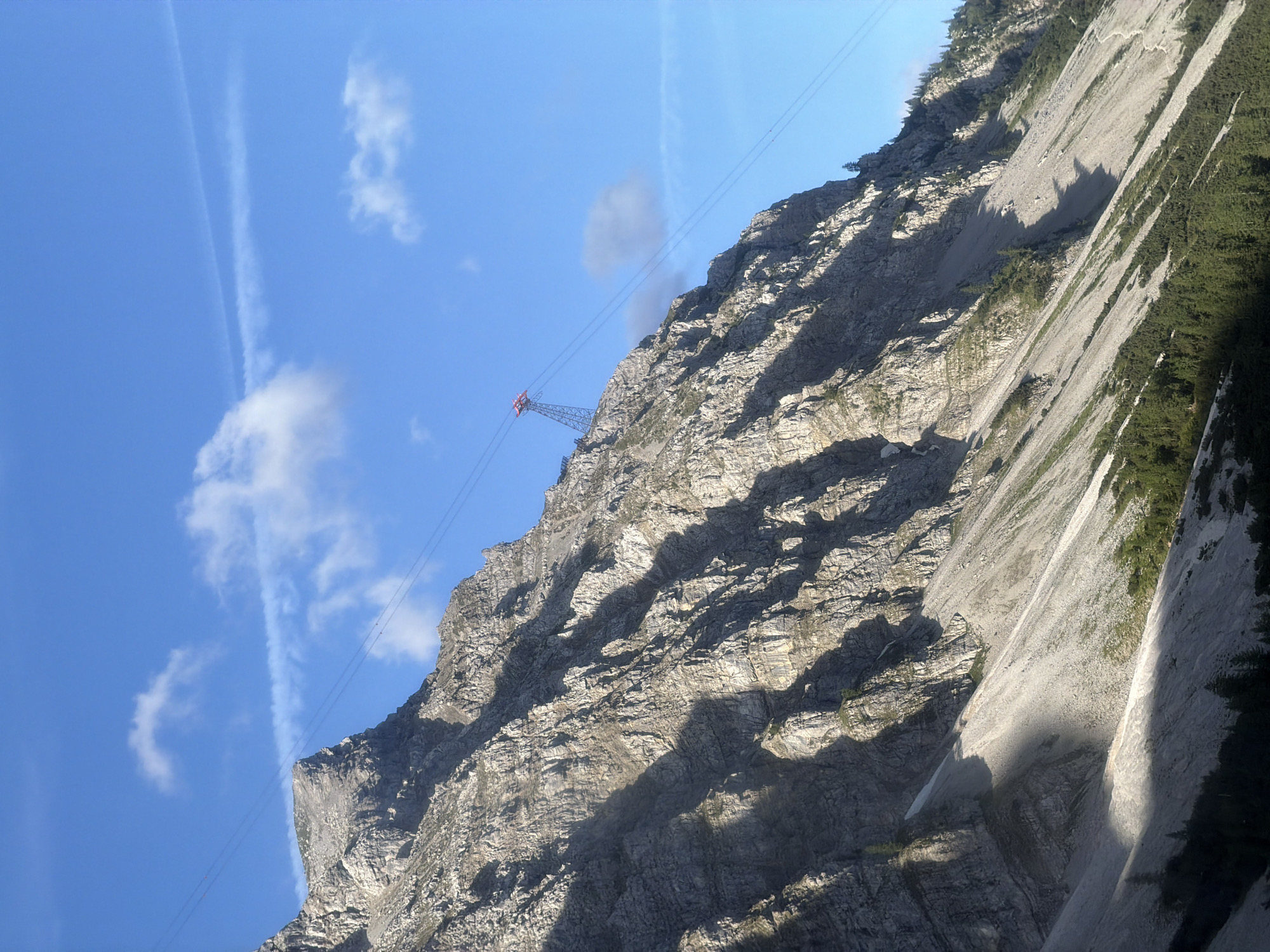 The image shows a steep, rocky mountain face in Grainau, Germany. A cable car ascends the mountain, with its red station visible at the top. The sky is a clear blue with some wispy clouds. The mountain is covered in scree and sparse vegetation. The sun casts long shadows across the mountainside, indicating it is either early morning or late afternoon.