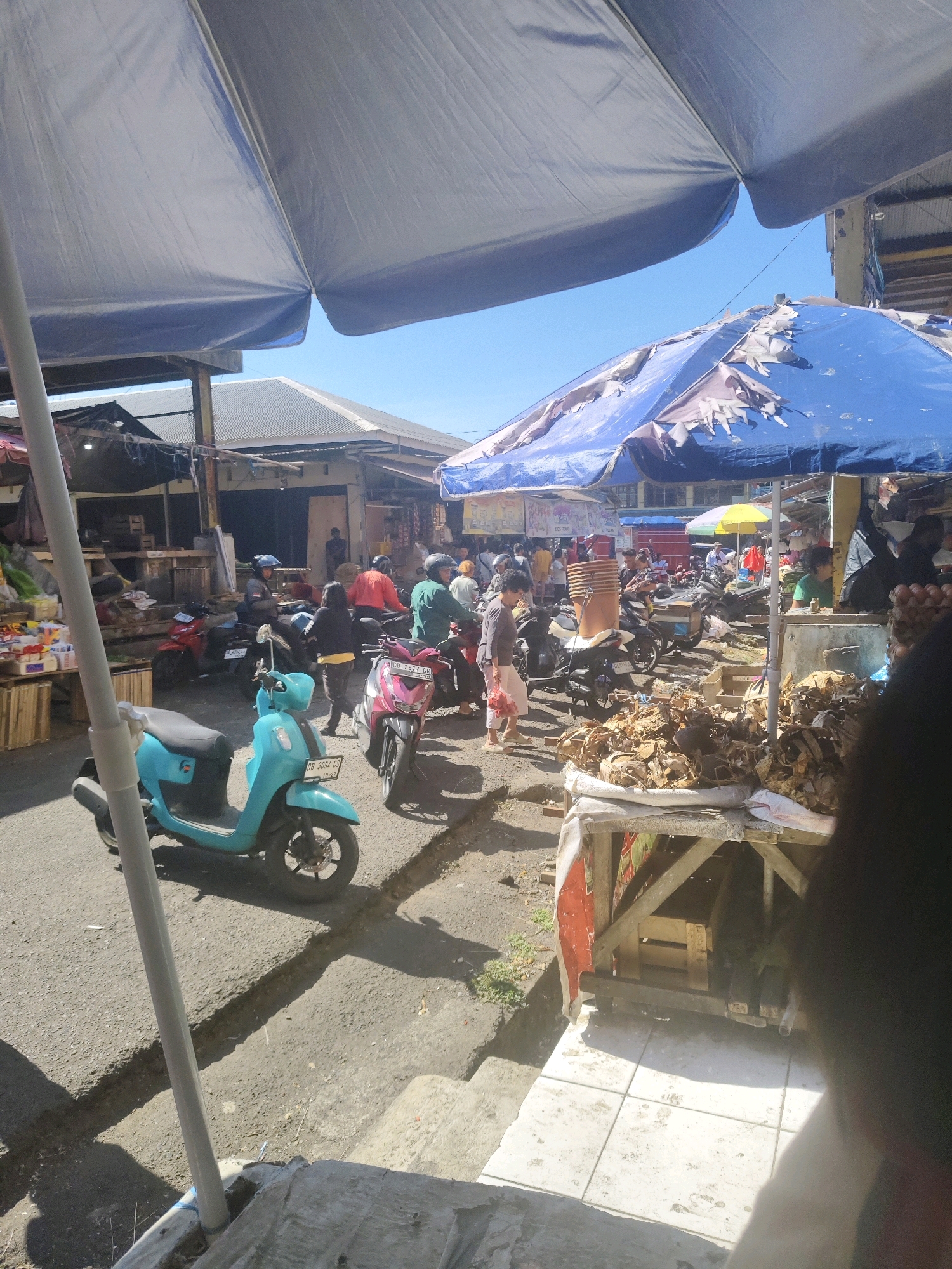 This is a street-level, eye-level, full shot of a bustling outdoor market scene under a bright blue sky, likely in Tomohon, Indonesia. The foreground is dominated by a large, light-colored umbrella, partially obscuring the view of the sky and the market stalls beyond. A thin, gray support pole for the umbrella is visible on the left side.
The market itself is a lively place, with a variety of people and vehicles. Several motorbikes are parked or in motion on the asphalt ground. A bright turquoise scooter is parked prominently in the left-middle of the frame, facing left. A pink and black motorbike is parked to its right. Various market stalls are set up, some covered with blue tarps, one of which appears weathered and torn. Stacks of goods, possibly produce or other market wares, are visible on wooden tables and crates.
Numerous individuals are scattered throughout the scene, engaged in activities such as shopping, selling, or simply moving through the market. Some are wearing helmets, suggesting they are riding or about to ride motorbikes. The overall impression is one of a busy, everyday market day. The architecture of the buildings in the background suggests a commercial area with modest structures. The ground is paved with asphalt and some areas show concrete or tiled surfaces. The lighting suggests it is daytime, with strong sunlight creating shadows.