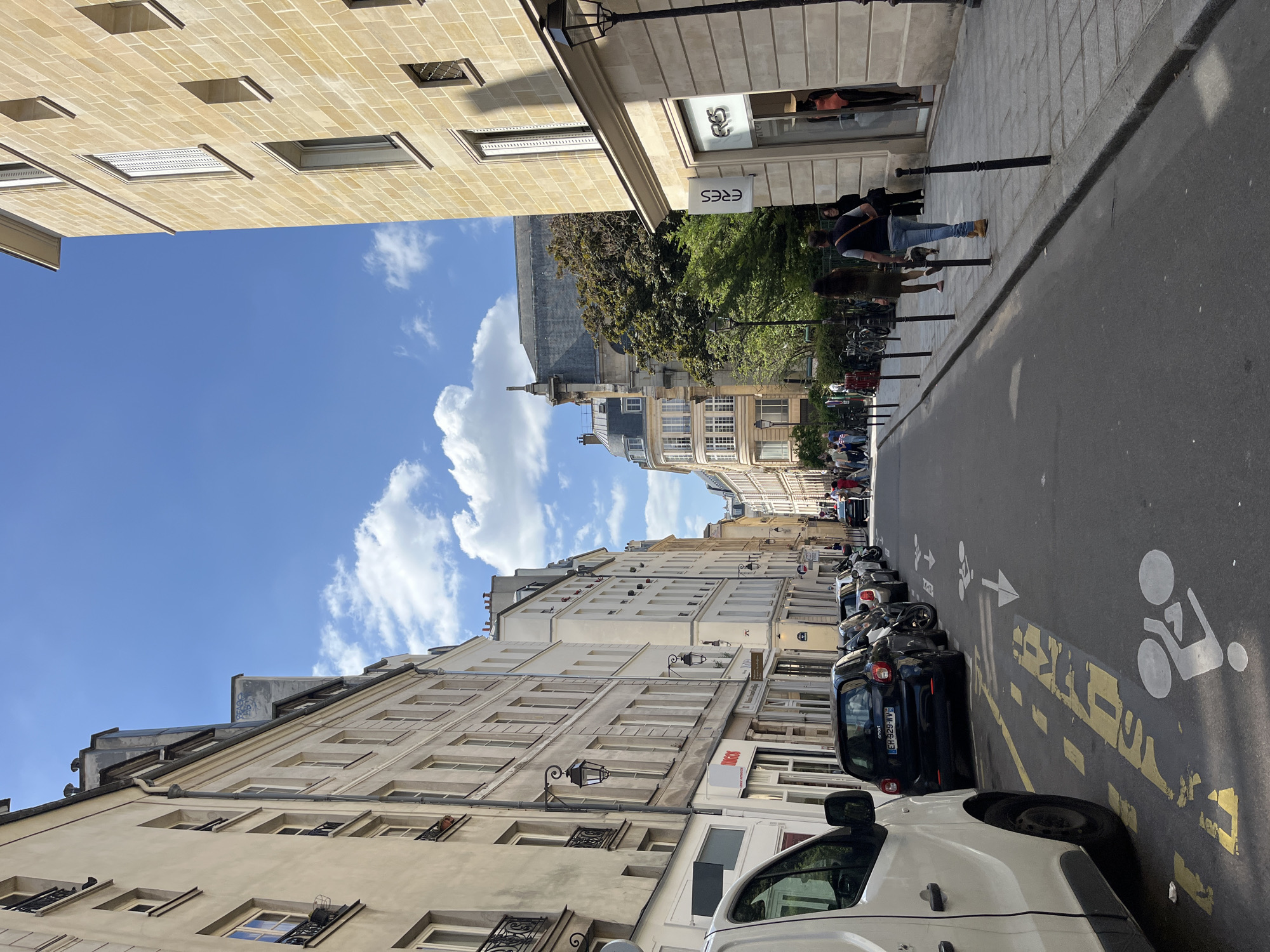 The image captures a street in Paris, France, showing a mix of architectural styles and typical Parisian urban elements.
The street is lined with buildings that feature light-colored facades and multiple windows, some with decorative ironwork. These buildings have varying heights and architectural details, reflecting a blend of styles over time. At least one building houses a store named "ERES".
The street itself is asphalt and contains a designated bike lane with painted symbols. Cars and motorcycles are parked along one side of the street, exhibiting the common tight parking conditions in urban areas.
Several people are walking on the sidewalk along the buildings, suggesting everyday activity in a residential or commercial area. The sky above is blue with scattered white clouds, creating a pleasant atmosphere.
