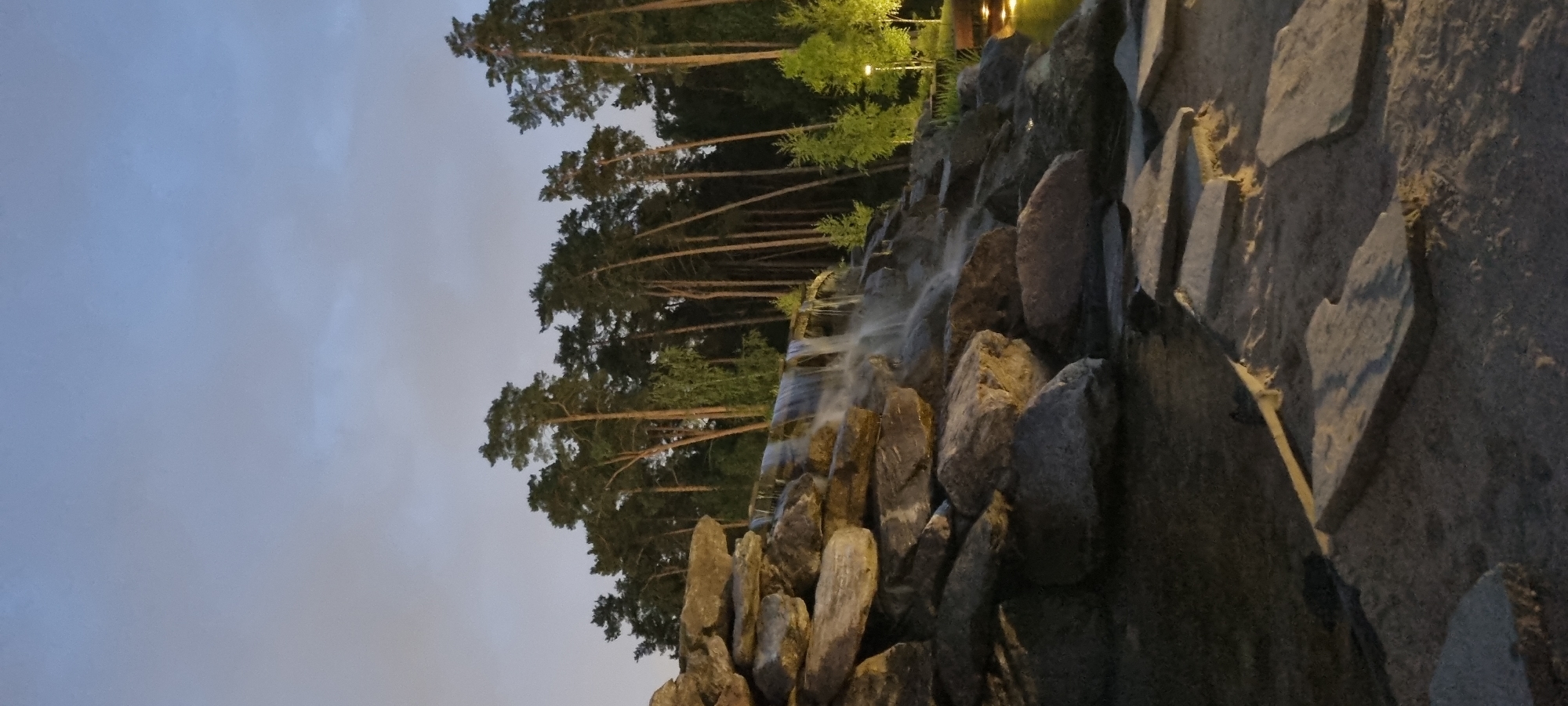 A man-made waterfall cascades over a pile of large, grey rocks. The waterfall is illuminated by artificial lights, creating a soft glow on the water. In the background, a dense forest of tall, slender pine trees stands against a dusky, pale blue sky. The ground in the foreground is covered with sand and large, flat grey stones arranged as stepping stones. To the right of the waterfall, the edge of a body of water with lights reflecting on its surface is visible. The scene is likely a park or garden area, possibly in Shulgino, Russia, given the pine trees and the style of landscaping.