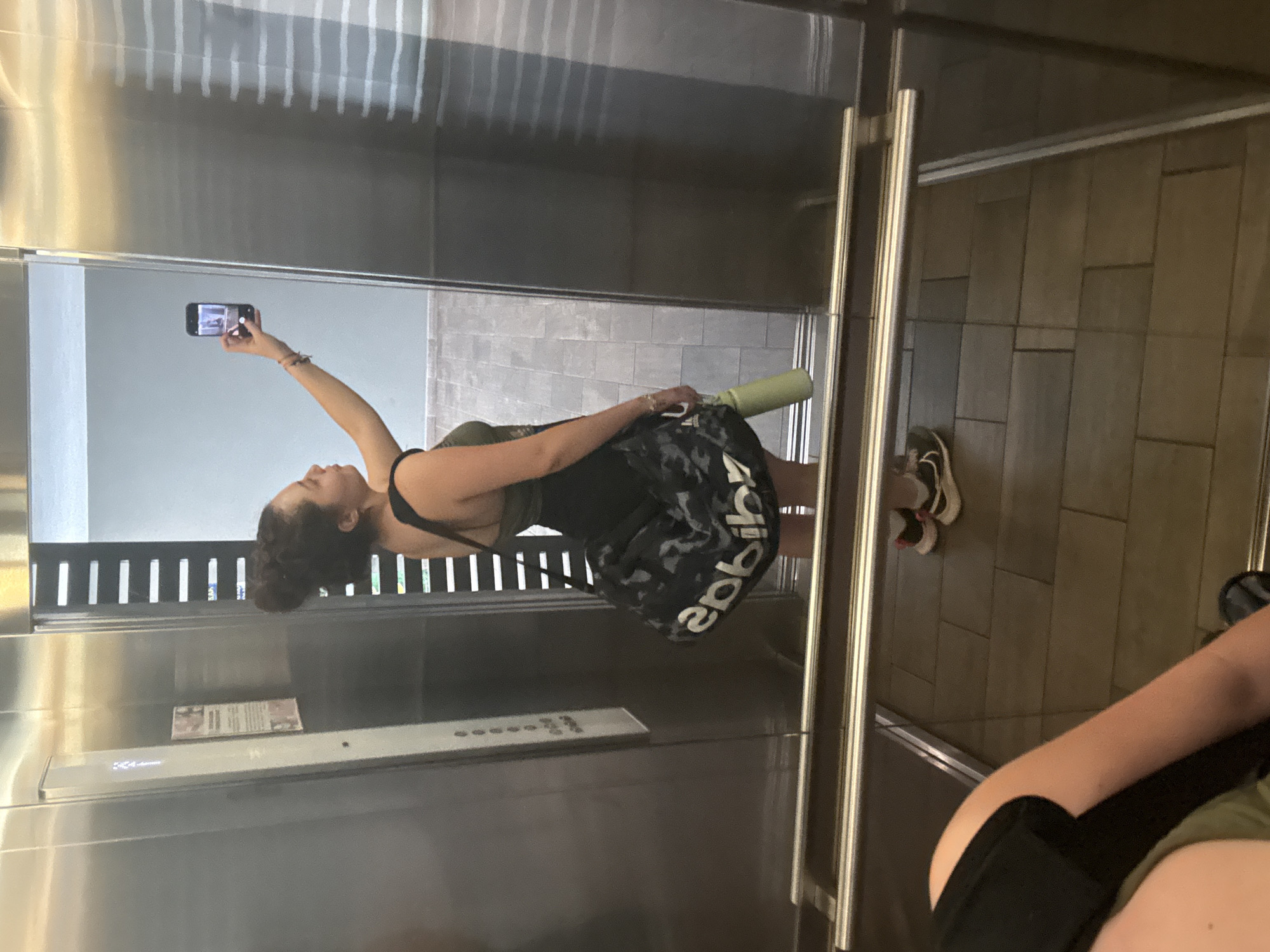 This image captures a mirror selfie taken by a woman inside an elevator, which is part of a building in Mexico City, Mexico. The perspective is from the woman’s smartphone, held in her right hand, pointed towards the reflective elevator door. The image has been rotated to show the scene upright.
The main subject is a young woman, likely in her 20s or 30s, with dark, curly hair pulled back. She is wearing athletic attire, including a dark-colored sports top or tank top and camouflage-patterned shorts or leggings. The word "adidas" is visible in white lettering on her left leg, reversed due to the reflection. In her left hand, she holds a light green water bottle.
The elevator interior features shiny, reflective walls, likely stainless steel. A control panel with illuminated buttons is visible on the left wall, with a '1' clearly displayed, possibly indicating the current floor. There's also a small notice or sign attached to the wall, though its text is illegible. The elevator doors are open, revealing a tiled floor and a modern building interior or hallway beyond, characterized by vertical slats and light-colored walls. A pair of sneakers (white with dark and possibly red accents) is visible on the floor, reflected near the bottom right. A partial reflection of another person's arm and torso, also wearing athletic-like clothing (green top and black bottom), is seen in the lower right corner, suggesting either the woman's own reflection from a different angle or another individual present in the elevator. The overall lighting suggests it is daytime.