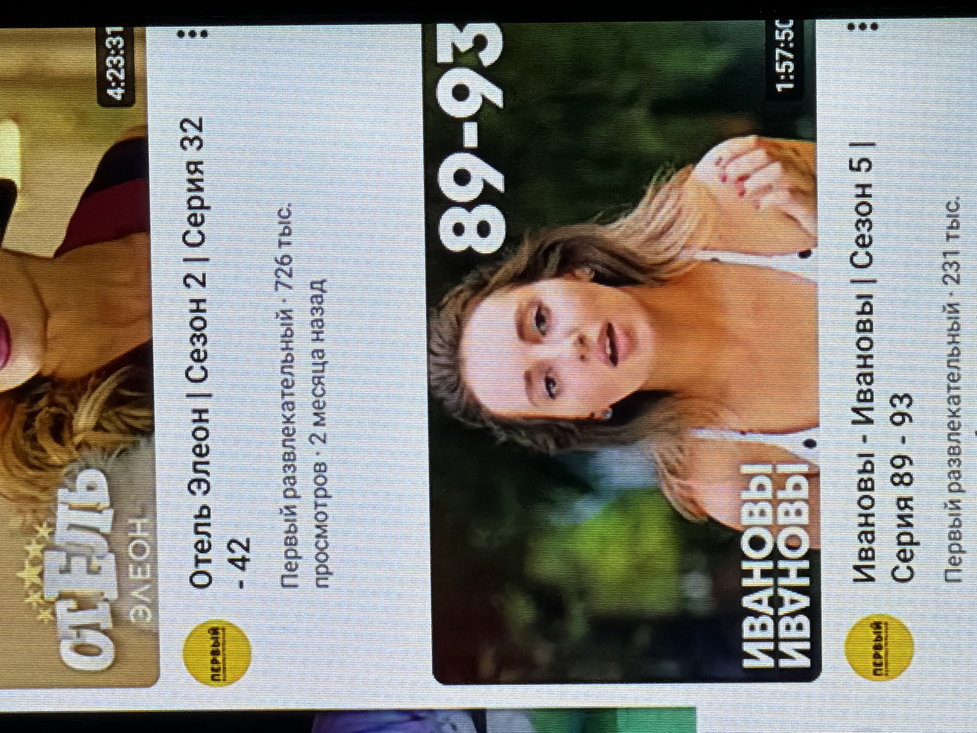The image shows a screen displaying content, likely from a video streaming service or a similar platform. Two distinct video thumbnails are visible, each with associated text and a circular "ПЕРВЫЙ" (First) logo.
The left thumbnail features a woman's upper body and face, with the text "Отель Элеон" (Hotel Eleon) prominently displayed. Below this, it indicates "Сезон 2 | Серия 32" (Season 2 | Episode 32), followed by viewing statistics: "726 тыс. просмотров 2 месяца назад" (726 thousand views 2 months ago). A timestamp "4:23:31" is also visible in the top left corner.
The right thumbnail shows a close-up of a woman's face and upper body. Large white numbers "89-93" are superimposed on the image. The text below identifies the content as "Ивановы - Ивановы | Сезон 5 | Серия 89 - 93" (Ivanovs - Ivanovs | Season 5 | Episode 89 - 93). Similar to the left, viewing statistics are provided: "231 тыс. просмотров" (231 thousand views), and a timestamp "1:57:50" is visible in the top right.
The overall scene depicts a digital interface presenting entertainment content, with a focus on Russian-language television series. The backdrop for the thumbnails is blurred, but appears to be green and outdoor-like, suggesting natural scenery. The image itself is a photograph of a screen, indicated by the horizontal lines characteristic of older display technologies. The context of Beșalma, Moldova, suggests this screen is located within that region.