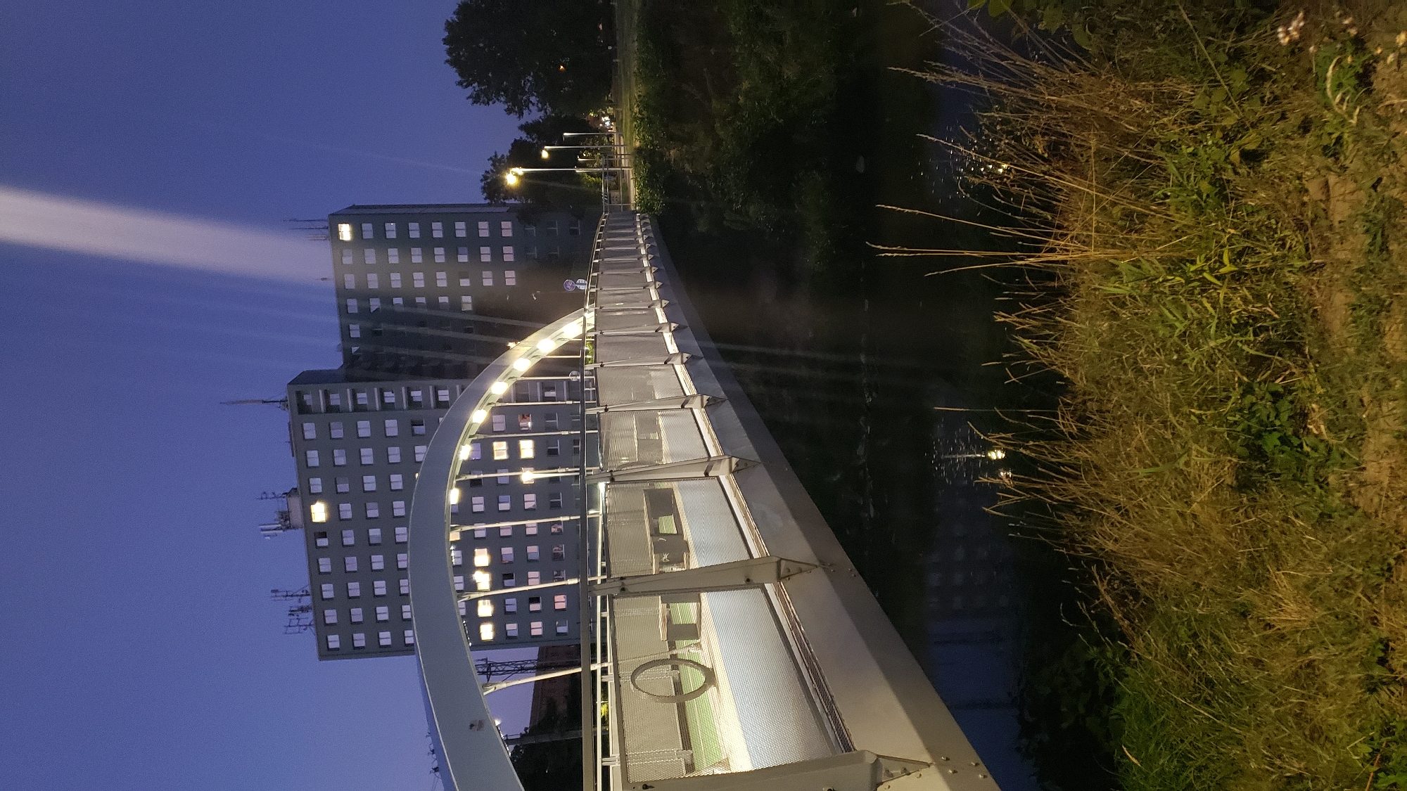 The image captures a modern pedestrian bridge spanning a body of water at twilight. The bridge features a sleek, curved design with illuminated railings and overhead lighting. On the bridge, there are benches and a circular decorative element. In the background, a tall, multi-story building with numerous windows, some of which are lit, dominates the skyline. Several antennas are visible on the roof of this building. A strong beam of light emanates from the sky behind the building, pointing downwards. Streetlights are visible along the path leading away from the bridge, and trees frame the scene on the right. The foreground consists of tall grasses and vegetation along the water's edge. The scene is characteristic of an urban environment, likely a residential area, at dusk or early evening. This pedestrian bridge is located in the Sekčov district of Prešov, Slovakia, specifically connecting the area near the elementary school on Šafárik Street to the residential blocks on Slánska cesta.