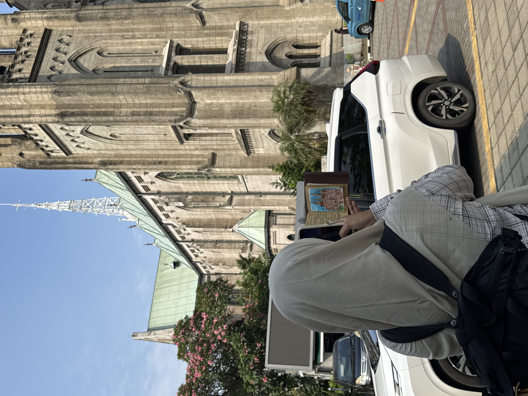 A person is photographing Jakarta Cathedral in Central Jakarta, Indonesia. The person is standing next to a white car on a paved area, with a backpack hanging on their shoulder. The cathedral's spires and intricate stone facade are visible in the background, partially obscured by trees and other vehicles.