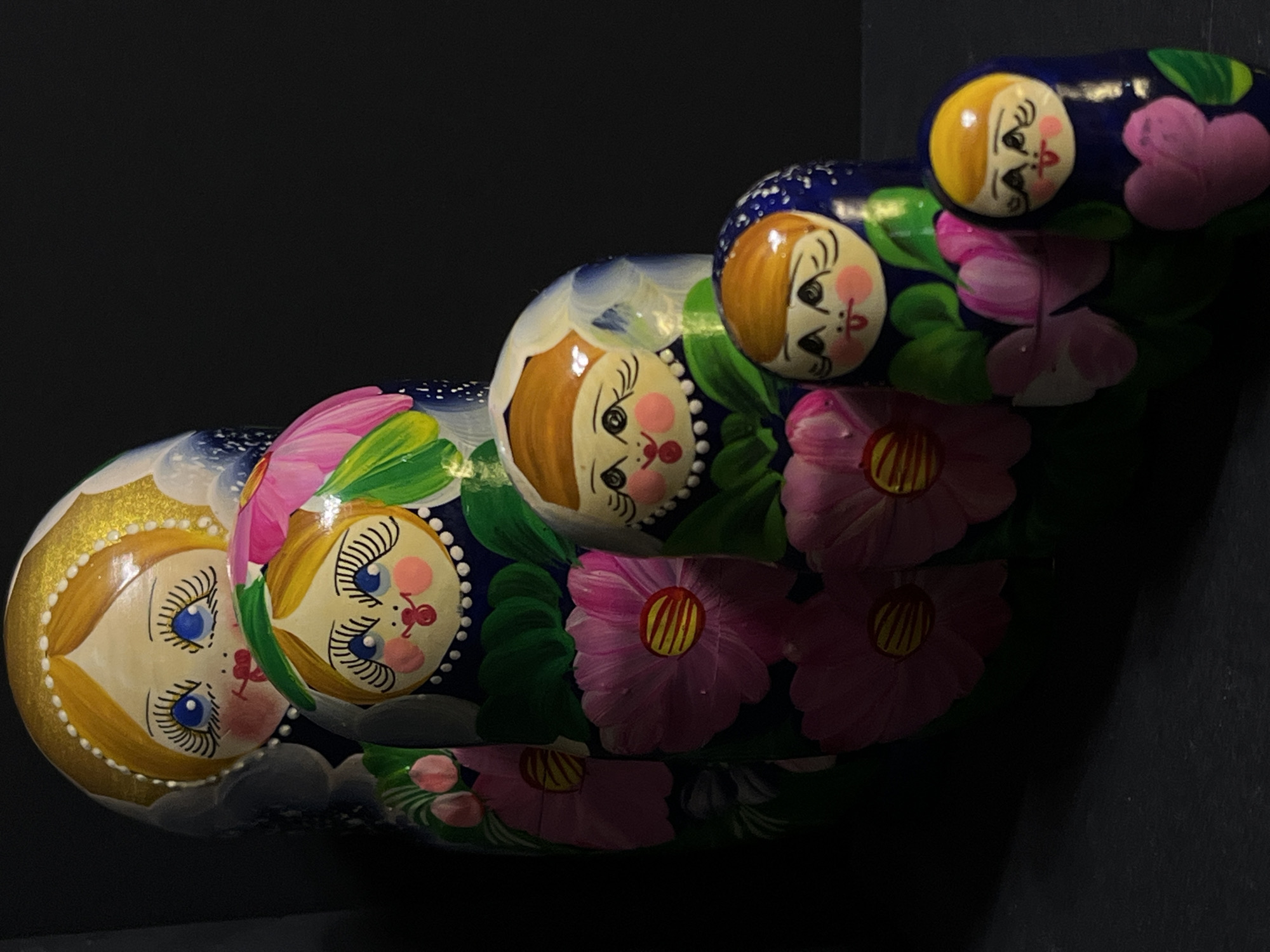 The image displays a set of six Russian nesting dolls, also known as Matryoshka dolls, arranged in a diagonal line against a dark, out-of-focus background. The dolls are progressively smaller, with the largest at the top left and the smallest at the bottom right. Each doll features a painted face with blue eyes, rosy cheeks, and red lips, set against a light skin tone. The dolls are adorned with floral patterns, predominantly large pink and green flowers. The largest doll has a golden headdress with pearl-like accents. The dolls are positioned on a dark, textured surface, possibly a table or floor. The lighting appears to be artificial, casting shadows and highlighting the glossy finish of the dolls. The scene is devoid of any human presence or activity. The image does not contain any specific geographical indicators that would allow for localization to Krasnyye Barrikady, Russia.
