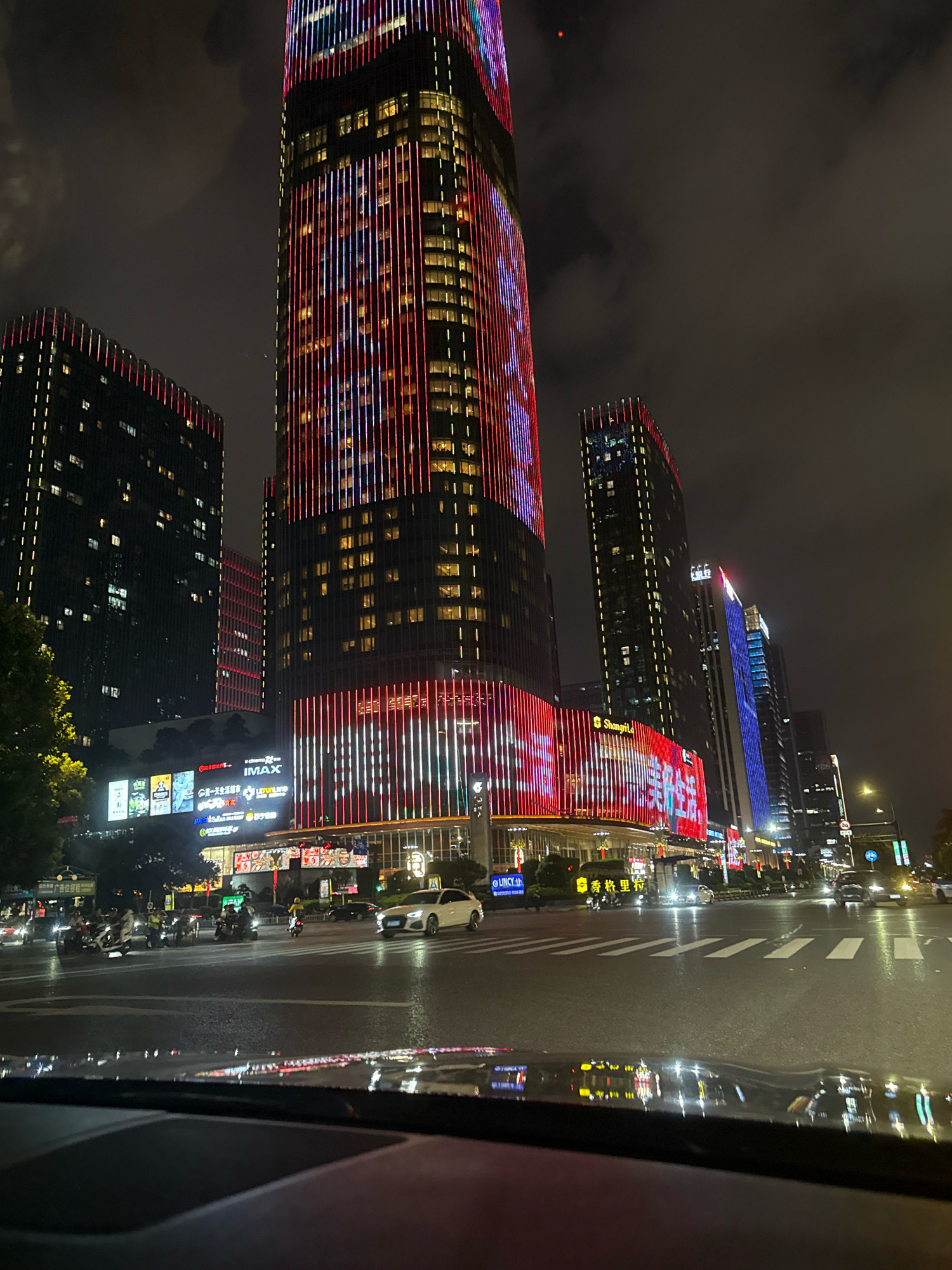 The image depicts a nighttime street scene in Yiwu, China, a city renowned for its wholesale markets. The foreground shows the interior of a vehicle, with the dashboard and windshield reflecting the illuminated cityscape. The main focus is on a cluster of modern skyscrapers and commercial buildings, brightly lit with colorful LED lights.
The tallest building on the left is adorned with vertical lines of red and blue lights, creating a dynamic visual effect. To its right, another building features similar vertical lighting in red and white. The lower sections of these buildings appear to house commercial establishments, indicated by illuminated signage, including "IMAX" and other Chinese characters that suggest shops and entertainment venues.
In the middle ground, a street is visible, wet from recent rain, with traffic consisting of several motorcycles, a white sedan, and other vehicles. Pedestrians are also present, mingling on the sidewalks and crossing the street. The traffic lights and street lamps illuminate the intersection, casting reflections on the wet asphalt. The overall atmosphere is one of a bustling, modern urban environment at night, characteristic of a major commercial hub like Yiwu. The sky is dark and cloudy, typical of an evening in a city.