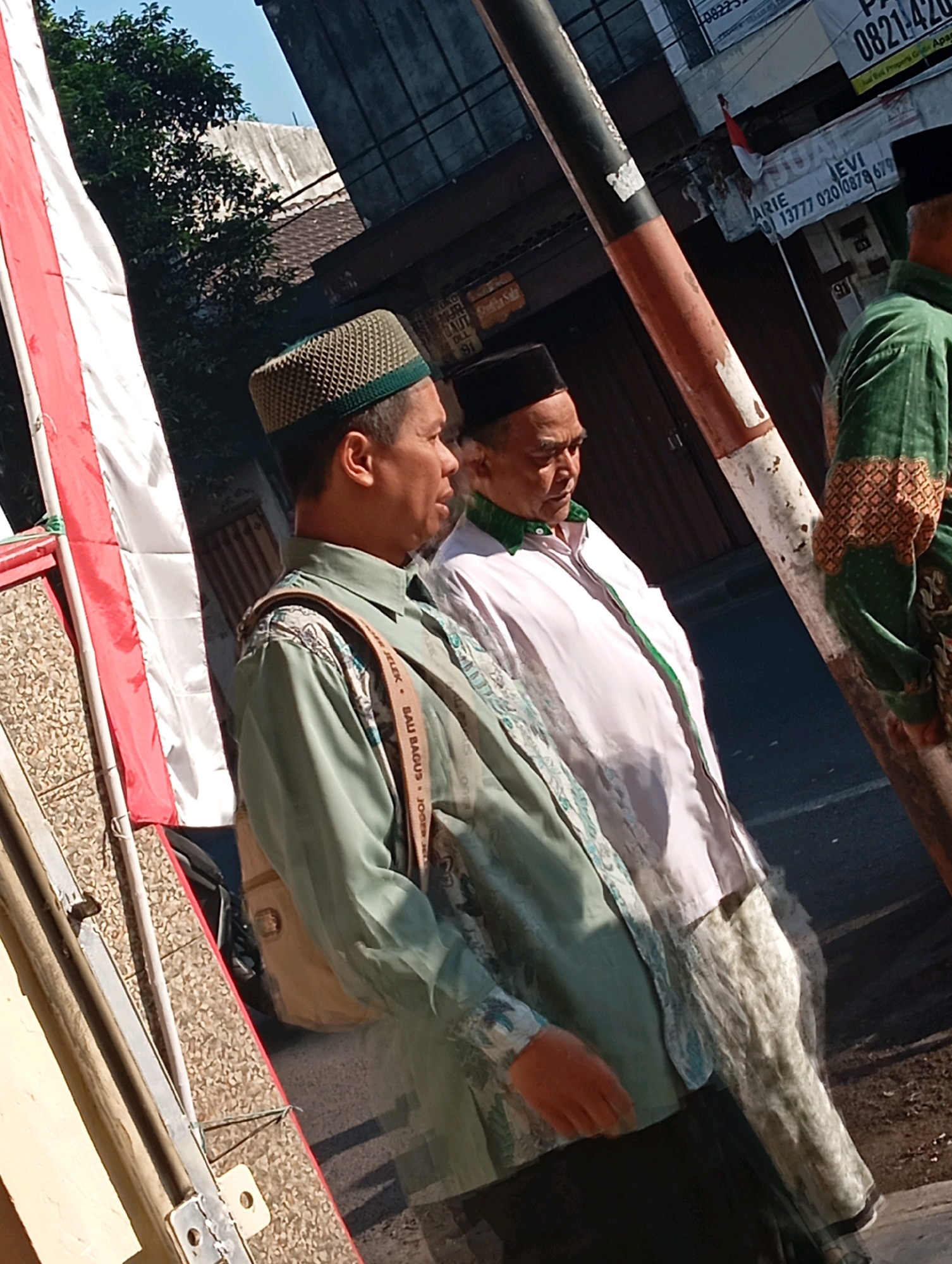 Two men, appearing to be of Southeast Asian descent and dressed in traditional Islamic attire, stand on a street in Malang, Indonesia. The man on the left wears a light green patterned shirt and a woven peci hat. He has a bag slung over his shoulder. The man on the right wears a white shirt with green trim and a black peci hat. He is partially obscured by a red and white pole. In the background, a building with a large dark doorway and a sign with Indonesian text is visible. A red and white flag, possibly the Indonesian flag, hangs from a pole on the left side of the image. The ground is paved and appears to be asphalt. The lighting suggests it is daytime, with sunlight casting shadows.
