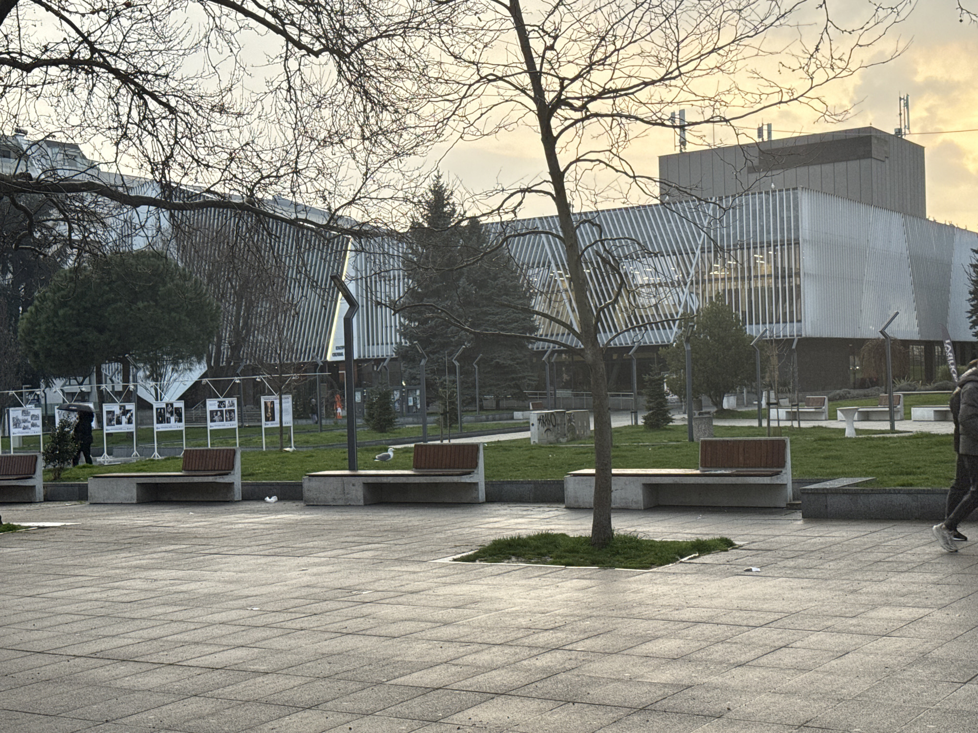 The image shows a plaza in Burgas, Bulgaria. In the background is the "Opera Burgas" building, characterized by its modern architectural style with vertical lines. In the foreground, there is a paved area with several benches. There are some trees in the middle ground. A few people are visible: one holding an umbrella, another is walking on the right side of the image. Display boards with photographs are located on the left. The weather appears overcast.