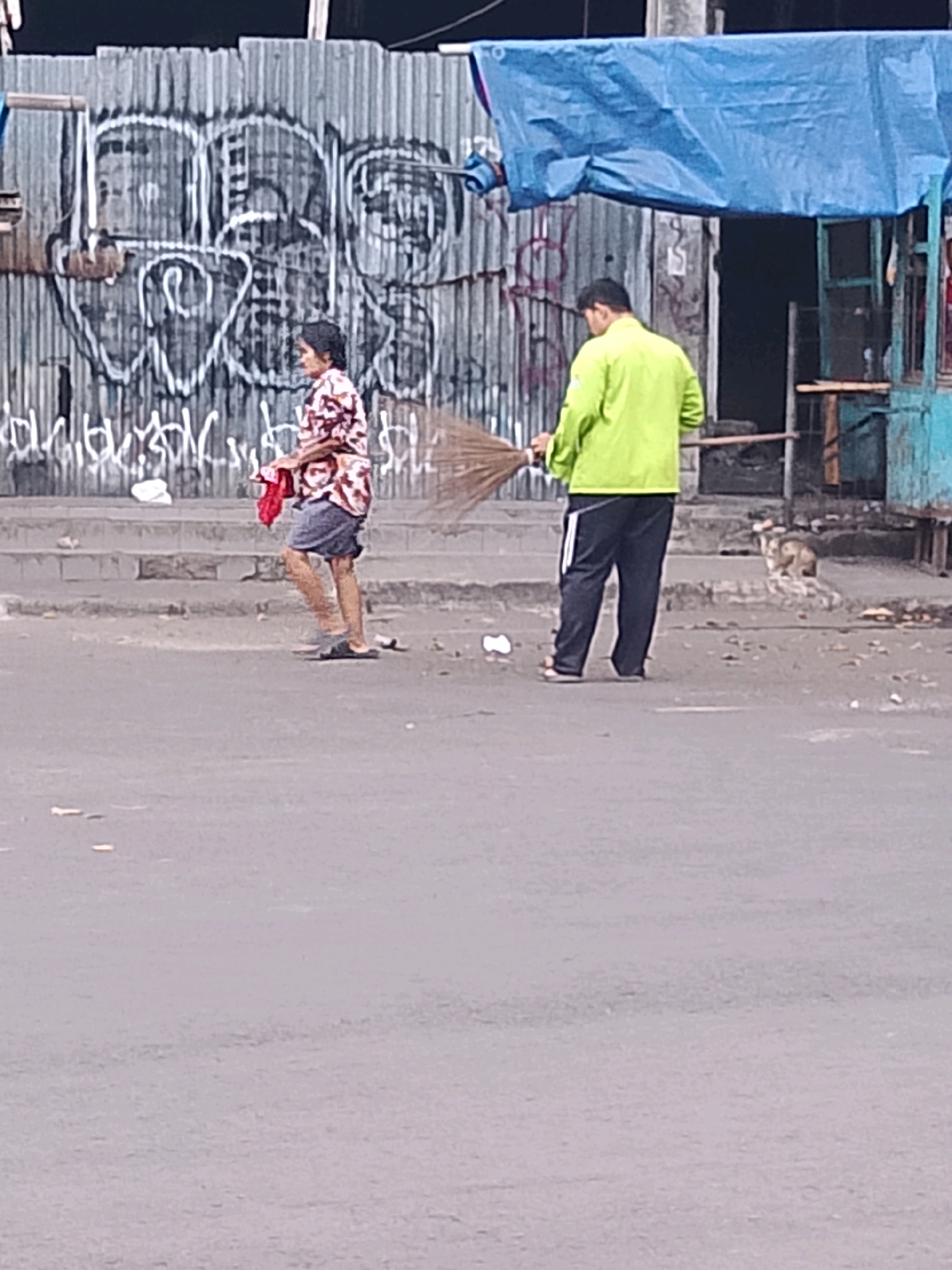 The image captures a street scene in Cirebon, Indonesia, with two individuals and a cat. On the left, a person in a patterned shirt and shorts walks with a red cloth in hand. To the right, another person, wearing a bright green long-sleeved shirt and dark pants, sweeps the ground with a broom. A cat is visible in the background, near a structure with a blue tarp overhead. The environment appears to be urban, with a corrugated metal wall covered in graffiti behind the individuals. The ground is paved, and debris is scattered around. The overall lighting suggests it is daytime.