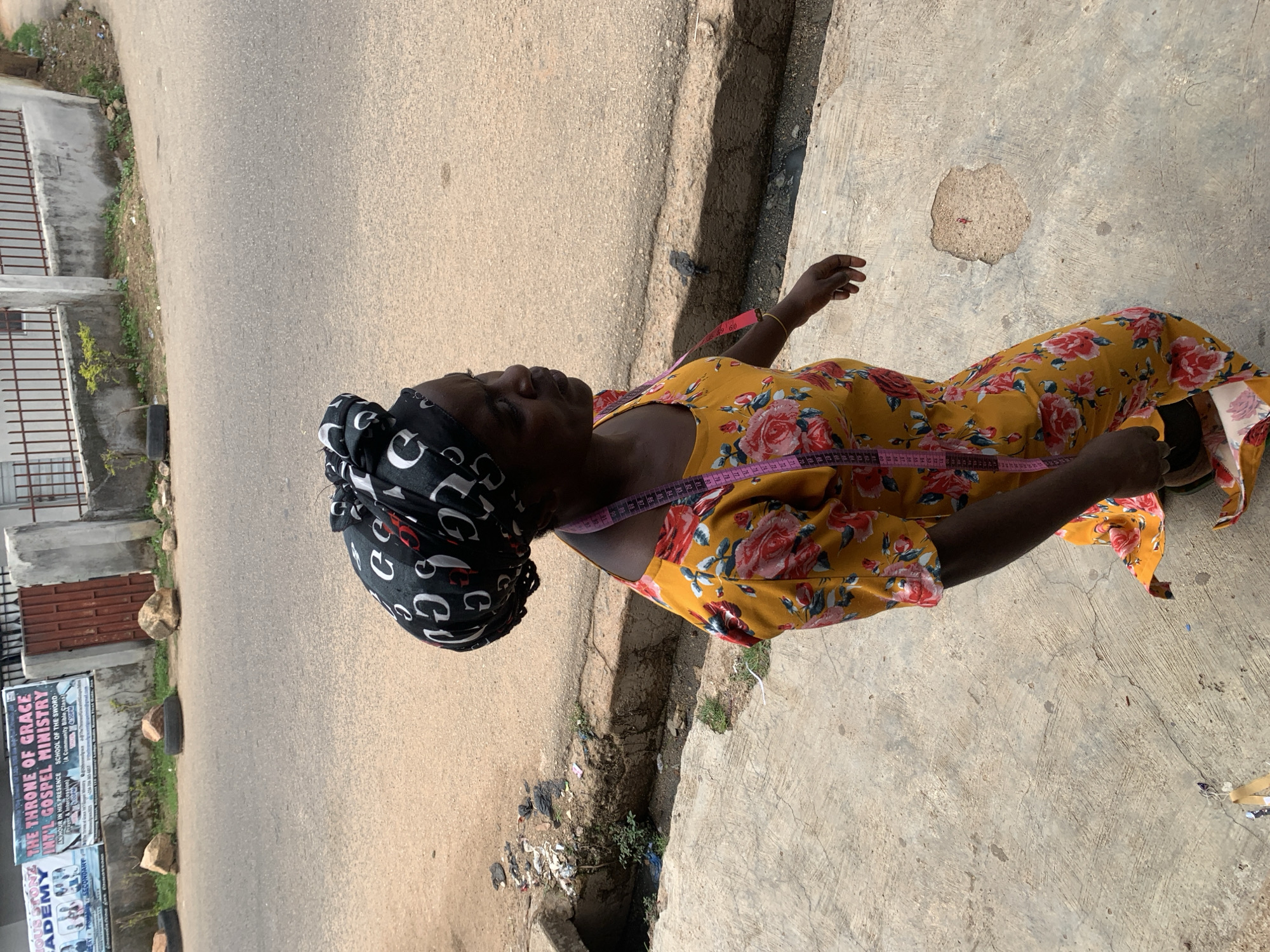 The image depicts an outdoor scene in Jos, Nigeria, featuring a dark-skinned adult woman in profile, walking on a concrete sidewalk beside an asphalt road. She wears a black headwrap patterned with white "G" letters and a vibrant yellow dress adorned with a floral print of red and pink roses and green leaves. A pink measuring tape with black markings is draped around her neck. Her right arm is slightly bent, and she looks to her right, seemingly observing the ground or the immediate vicinity.
The concrete sidewalk beneath her is worn, with visible cracks and some exposed gravel, including a small, reddish patch. To the immediate left of the sidewalk is a concrete-lined drainage gutter containing some litter and sparse green vegetation. Beyond the gutter, the asphalt road shows signs of wear and minor cracks.
The background features a low concrete wall with a brown metal fence on top, enclosing what appears to be a property. A brown metal gate is visible further down the wall. Along the side of the road, several large rocks and at least two discarded black tires are present, some appearing to act as informal barriers. Above the wall, two large banners are affixed: the left banner partially reads "...IOUS STONE ACADEMY," while the right banner clearly states "THE THRONE OF GRACE INTL GOSPEL MINISTRY," with additional text including "AN HOUR IN HIS PRESENCE," "SCHOOL OF THE SWORD," and "A Community Bible Class." Small patches of dry earth and sparse greenery are visible between the road and the wall. The scene is illuminated by natural daylight, casting soft shadows.