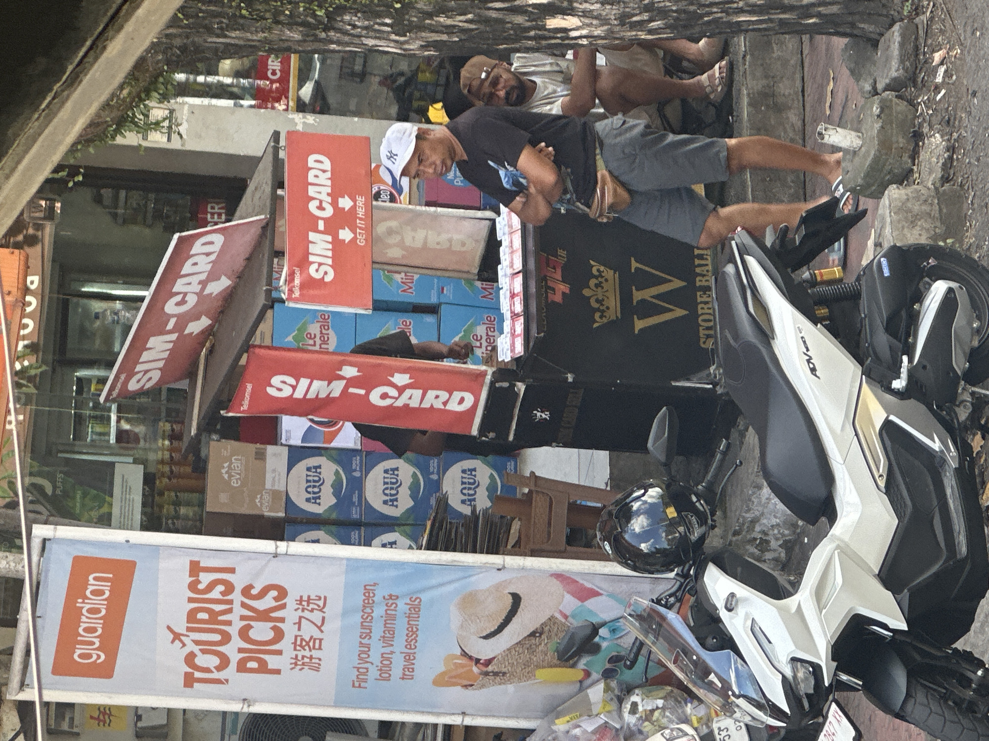 In Ubud District, Indonesia, outside a store, a white motorcycle is parked in the foreground. Behind it, a street vendor stall displays SIM cards. A man wearing a white cap and black t-shirt, with a brown fanny pack, stands with his arms crossed. Another man sits behind him. The stall has signage for "SIM-CARD" and "STORE BALI", along with stacks of "AQUA" bottled water. A tall vertical banner advertising "GUARDIAN TOURIST PICKS" with travel essentials is visible to the left. The environment appears to be an outdoor market or street scene.