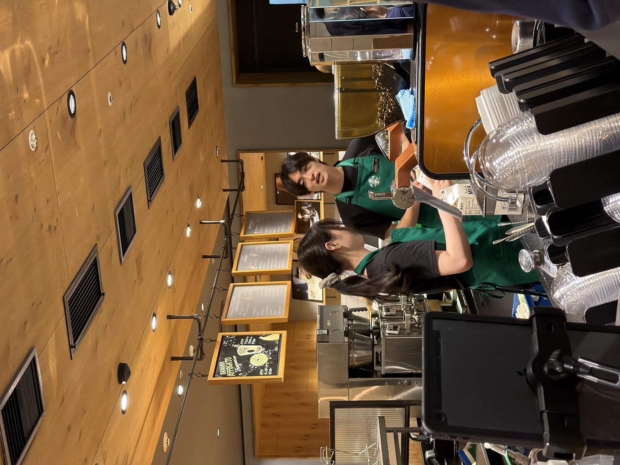 This image shows the interior of a Starbucks located in Chuo, Japan. Two baristas, a man and a woman, are visible behind a counter, engaged in work. The male barista, wearing a green apron over a black t-shirt, is speaking while looking towards the woman. The female barista, also in a green apron over a black t-shirt, has her hair in a ponytail and is looking down. In front of them are various coffee-making equipment, including a large espresso machine and a grinder filled with coffee beans. Stacked clear plastic cups and white paper cups are also present on the counter, along with a carton of milk and other service items. The environment features a wooden ceiling with modern lighting and ventilation fixtures, and menus are displayed on the wall behind the baristas.
