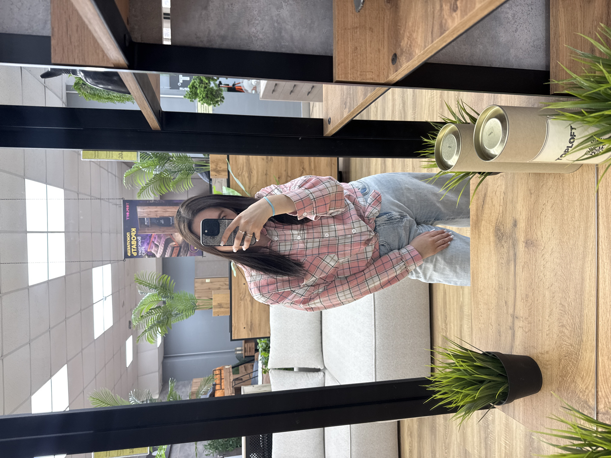 The image displays a mirror selfie taken indoors, likely within a furniture showroom in the city of Chaikovsky, Russia. The reflection shows a female individual with long, dark hair, holding a dark-colored smartphone to capture the image, which partially obscures her face. She is wearing a pink, white, and grey plaid long-sleeved shirt tied at the waist, light blue denim jeans, several rings on her left hand, and a light blue elastic bracelet on her right wrist.
The foreground directly in front of the mirror features a light wooden surface, possibly a console table or shelf. On this surface, to the left, is a small black pot containing artificial green grass-like plants. To the right, two vertical, cylindrical cardboard containers with metallic tops are present, one partially obscured, the visible one bearing a label with "TOPLOFT" text and a QR code, also surrounded by artificial green grass.
The environment reflected in the mirror depicts a commercial space with a light wood-look floor and a drop ceiling with white panels and recessed rectangular light fixtures. In the left background of the reflection, a light grey modular sofa composed of multiple sections with backrests is visible. Further back, artificial palm-like plants in wooden planters and various wooden display units or furniture pieces populate the area. Overhead signs, reflected and thus horizontally mirrored, feature Cyrillic text, including "КРОВАТИ" (Beds) and other furniture-related words, consistent with a furniture retail setting in Russia. To the immediate right of the mirror's frame, a dark-framed shelving unit with light wooden shelves is visible, holding a black decorative object on an upper shelf.
