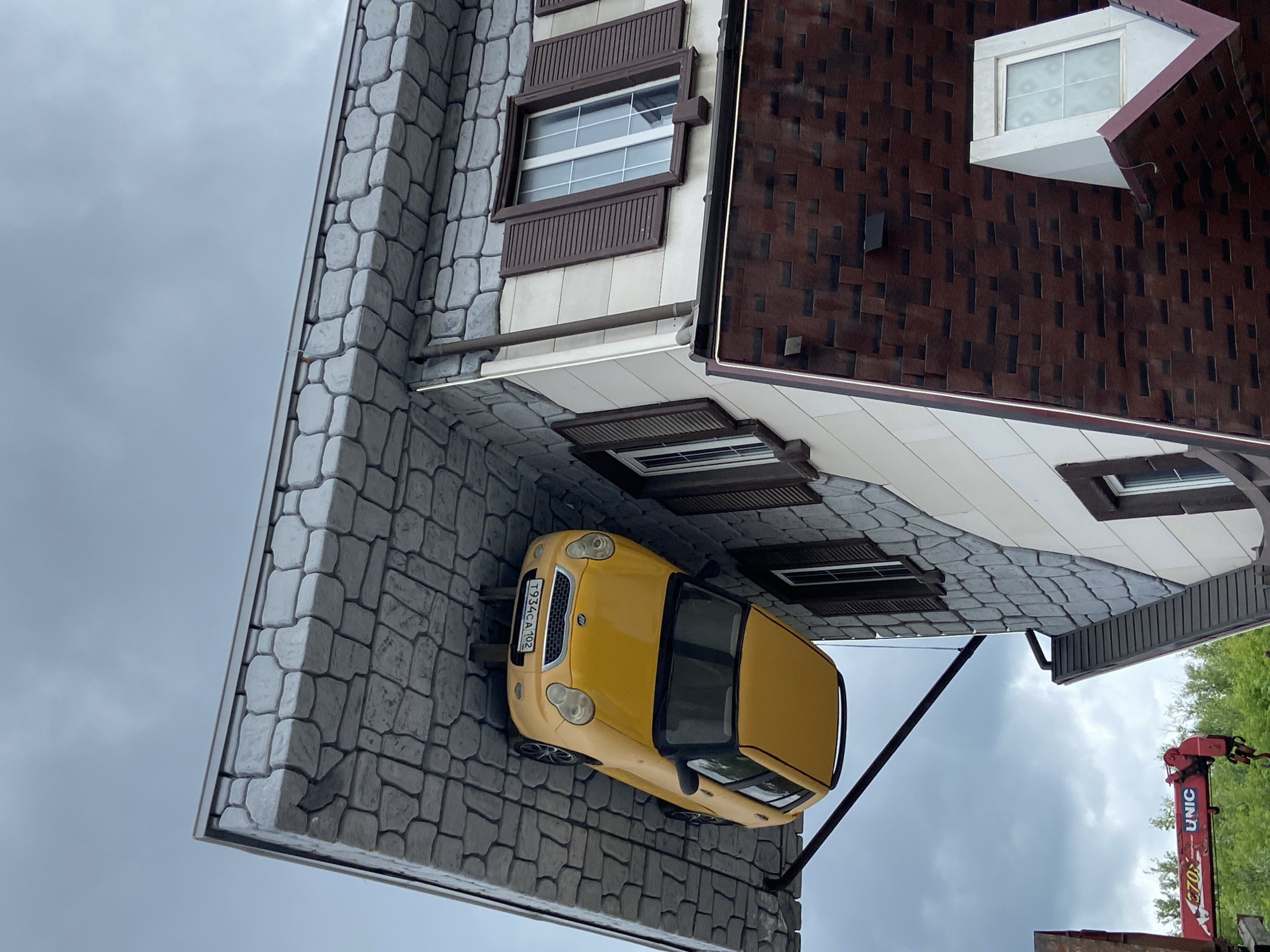 An upside-down yellow car is attached to the underside of a building's roof, which is made of gray stone-like material. The car's license plate reads "T934CA 102". The building has windows with brown shutters and a dark red shingled roof. In the background, a red crane with the logo "UNIC" is visible. The scene is set against a cloudy sky and appears to be an attraction or art installation. This structure is located at the "Upside Down House" (Dom Vverkh Nogami) in Ufa, Russia.