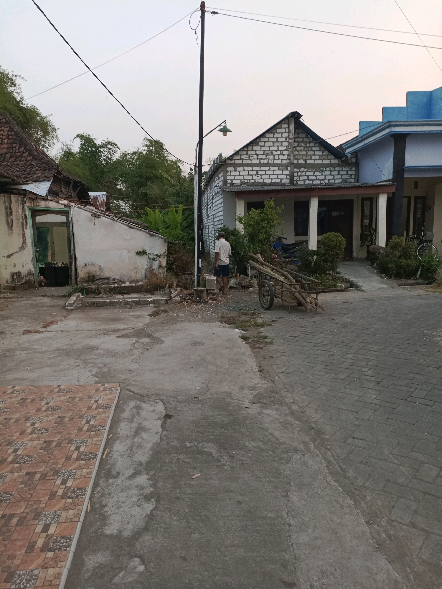In Rancang Kencono, Indonesia, an outdoor scene depicts a street with a man walking towards the left. The man is wearing a light-colored shirt and dark shorts. In front of him, a handcart is loaded with logs. To the right of the man and cart, a building with a light-colored brick facade and a dark roof stands, with several trees and bushes in front of it. To the left of the scene, a weathered, light-colored building with a damaged roof and an open doorway is visible, surrounded by overgrown vegetation. Overhead, power lines crisscross the sky, with a streetlamp attached to a pole on the right. The ground is a mix of concrete, paved stones, and dirt.