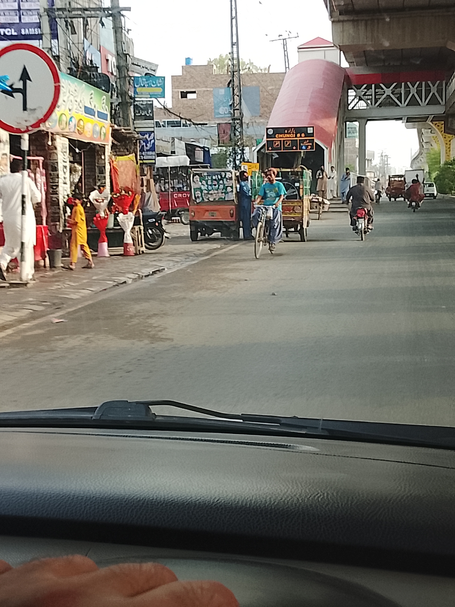 The image depicts a street scene in Gulgasht Colony, Pakistan. A road sign indicating a choice of directions stands at the corner of a street lined with shops and vendors. Several people are walking on the sidewalk, including one in a white robe and another in yellow clothing.
In the road, a man is riding a bicycle, and several motorized rickshaws and motorcycles are also present. A pedestrian overpass with a red arch is visible in the background. The image appears to be taken from inside a vehicle, with the dashboard and windshield wipers visible in the foreground.