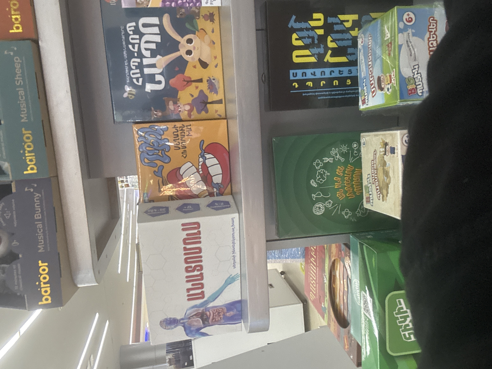 The media file presents an indoor view of a retail space, likely a toy or bookstore, with shelves laden with various boxed games and children's products. The setting appears to be in Yerevan, Armenia, as indicated by the predominant Armenian text on the product packaging and the word "երևան" (Yerevan) visible on one of the boxes towards the bottom right.
The shelves are light-colored, possibly metal or wood, and illuminated by bright overhead lighting. On the top left shelf, two items are visible: "baroor Musical Sheep" and "baroor Musical Bunny." Directly below these, a large white box features a detailed illustration of human anatomy and is labeled "ԱՆԱՏՈՄՈւՍ" (Anatomus), described as an "սեղանի ինտելեկտուալ խաղ" (intellectual board game) suitable for "12+" ages and "2-4" players, with a "30-40" minute playtime.
On the shelf above it, a dark blue box with a rabbit illustration reads "ՀՈՊ-ՀՈՊ" (Hop-Hop) and states "ԽԱՂՈՒՄ ԵՆ ԱՄԵՆԱՓՈՔՐԵՐԸ" (Played by the youngest ones). Adjacent to this, a black box is inscribed with "ՍՈՎՈՐԵՑ ԽԱՂ ԴՊՐՈՑ" (Learn, Play, School Game). Below the "Hop-Hop" game, an orange box displays a cartoonish mouth and partial text "ՊԱՏՄՈՒԹ" (likely 'History' or 'Story'). To the right, a small green and blue box is marked "6+" and "BIG ՊՍՏԻԿ" (Big Little) "ՄԱՐԶԱՁԵՎ" (Sport type/discipline). A green box below it features whimsical white line drawings.
A person's arm, dressed in a dark sleeve, is visible on the right side of the frame, partially obscuring some items and suggesting someone is actively browsing the selection. The overall impression is a well-stocked store offering educational and recreational items for children and families.