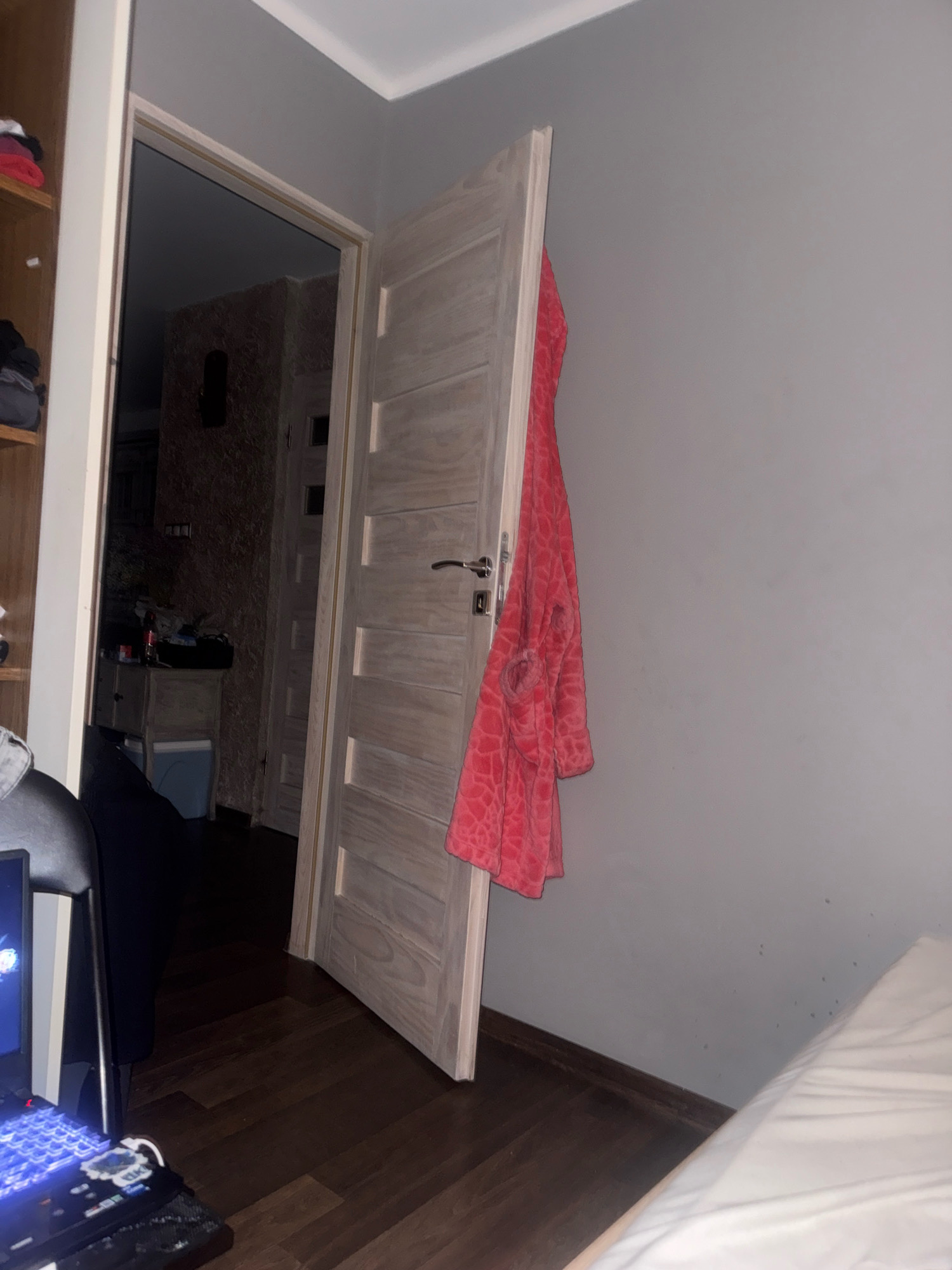 The image depicts an interior scene of a room, likely a bedroom or a hallway connecting to one. In the foreground, a wooden floor leads towards an open doorway. To the left, a portion of a shelf unit is visible, displaying folded clothing. A black chair and a laptop with a lit blue keyboard are also situated in the left side of the frame.
The doorway reveals a dimly lit hallway with patterned wallpaper. A light-colored wooden dresser with drawers and a light blue bin are visible within this hallway. Further down the hallway, another open doorway suggests another room or passage.
The primary focus of the image is a light wood-colored door that is open inwards, revealing its interior side with horizontal paneling and a silver door handle. A pink bathrobe with a textured pattern is draped over the edge of this open door. The walls of the room are painted a neutral gray. In the background, on the right side of the image, a portion of a white mattress is visible.
There is no information in the image to suggest a specific location in Gorzow, Poland.