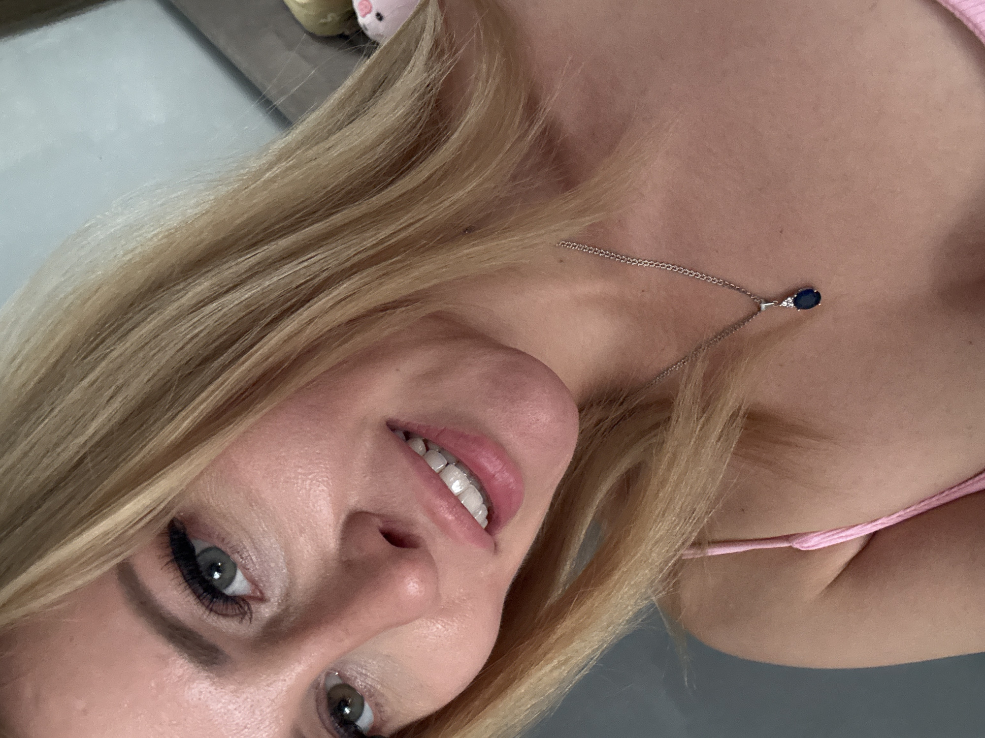 A close-up, slightly angled portrait of a blonde woman, likely in her 20s or 30s, smiling directly at the camera. She has fair skin, visible pores, and green/hazel eyes with prominent, dark eyelashes. Her mouth is open in a smile, revealing straight, white upper teeth, and her lips are pink. Her straight, blonde hair falls around her shoulders and chest. She is wearing a pink spaghetti strap top and a silver-toned chain necklace with a pendant featuring an oval, dark blue gemstone (likely sapphire) accented by two small clear stones (likely diamonds). The background is blurred, showing a light grey wall to the left and a lighter blue/grey wall or surface to the upper right, with indistinct shapes of objects, suggesting an indoor setting in Unknown, Unknown.